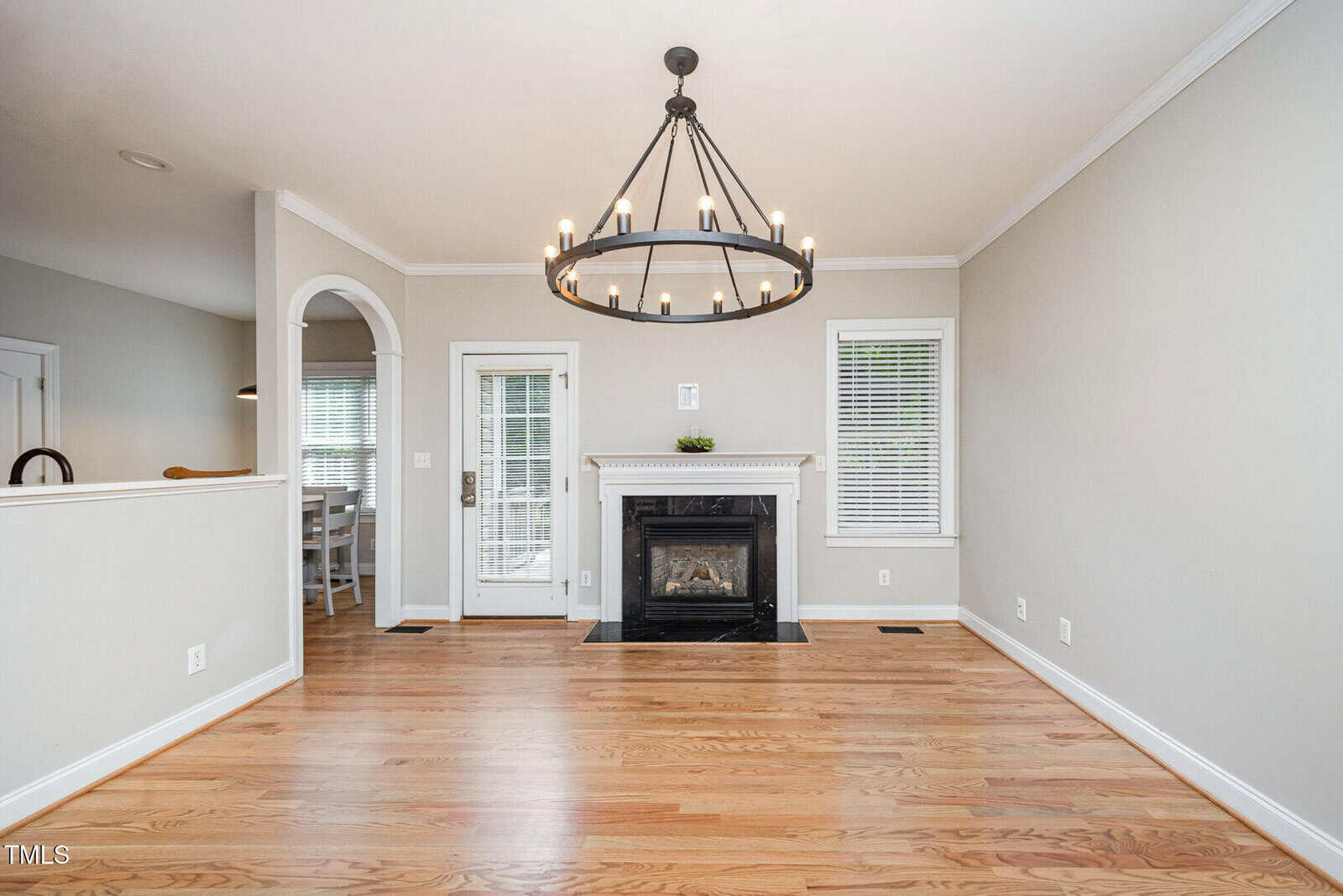 A living room with light wood floors, a black fireplace with a white mantel, a round black chandelier, two windows, and a glass door. Neutral walls, white trim, and an open archway lead to another room.