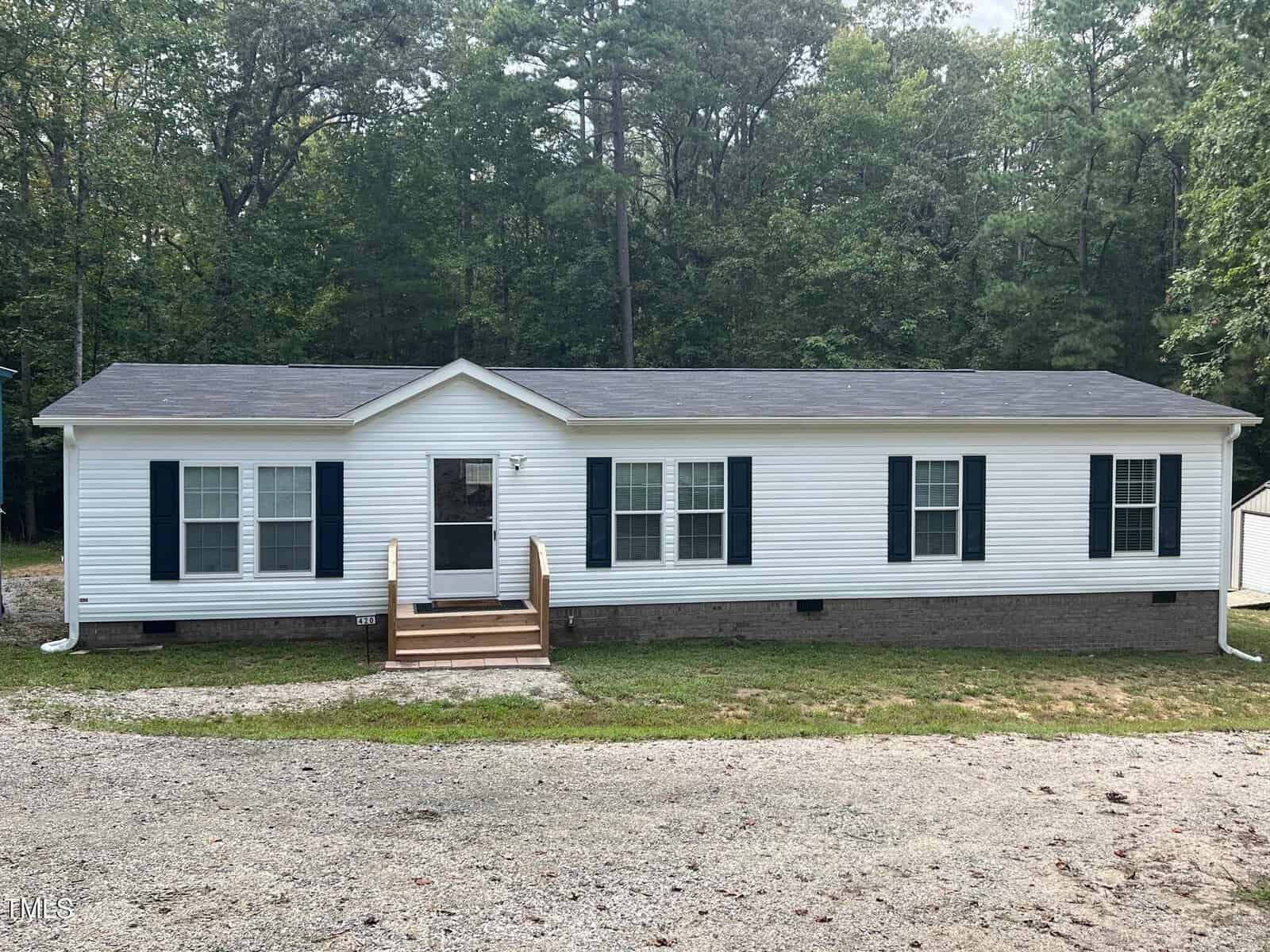 A single-story white manufactured home with black shutters, a dark roof, and a small front porch sits in front of a wooded area. The yard is gravel and grass, and the house has multiple windows along the front.