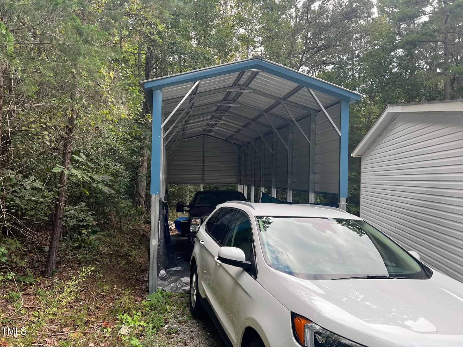 A white SUV is parked in front of a metal carport, which shelters another vehicle. The carport is next to a white building, surrounded by trees and greenery.