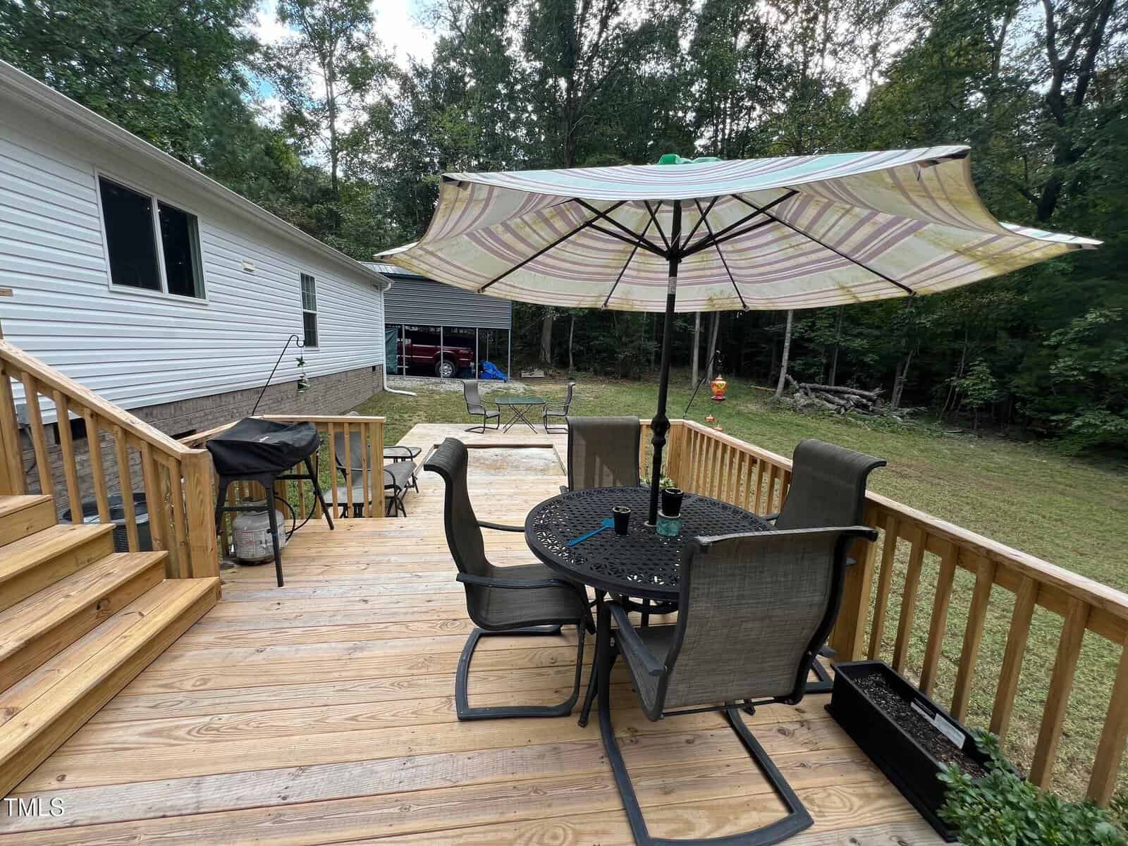 A wooden deck with a round table, four chairs, a large striped umbrella, and a grill beside wooden stairs. The deck overlooks a yard with trees, a shed, and outdoor furniture in the background.