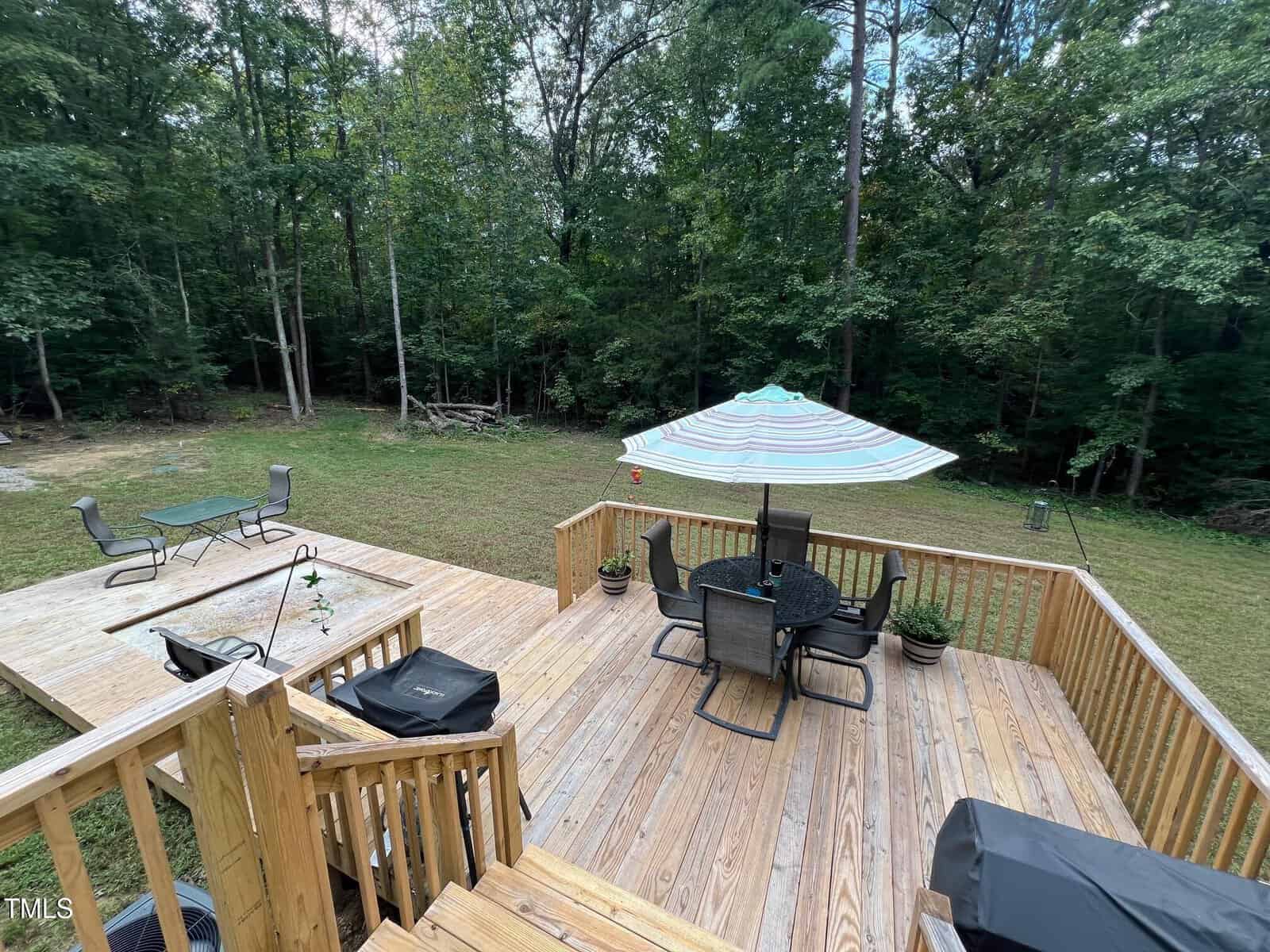 A wooden deck with a table, chairs, and a large striped umbrella overlooks a grassy yard bordered by dense trees. An additional seating area with chairs and a table is visible on a lower platform.