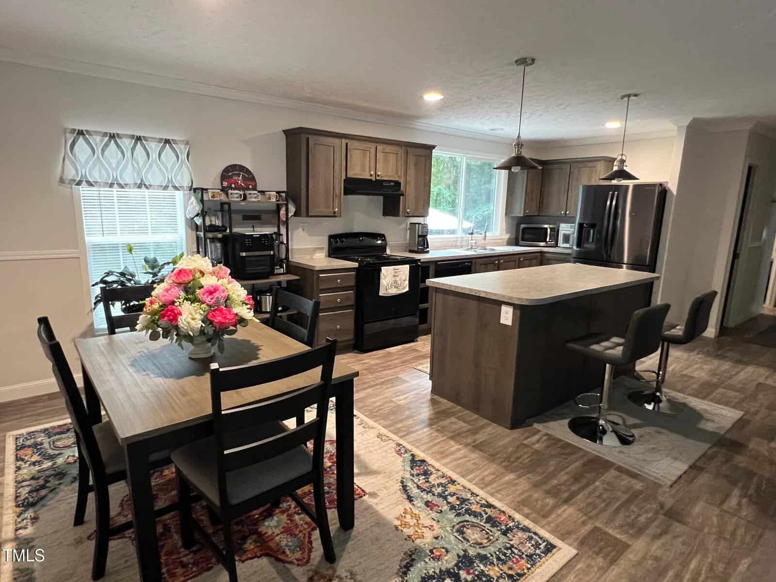 A modern kitchen and dining area with wooden cabinets, a central island with two barstools, a dining table with six chairs and a flower arrangement, stainless steel appliances, and large windows letting in natural light.