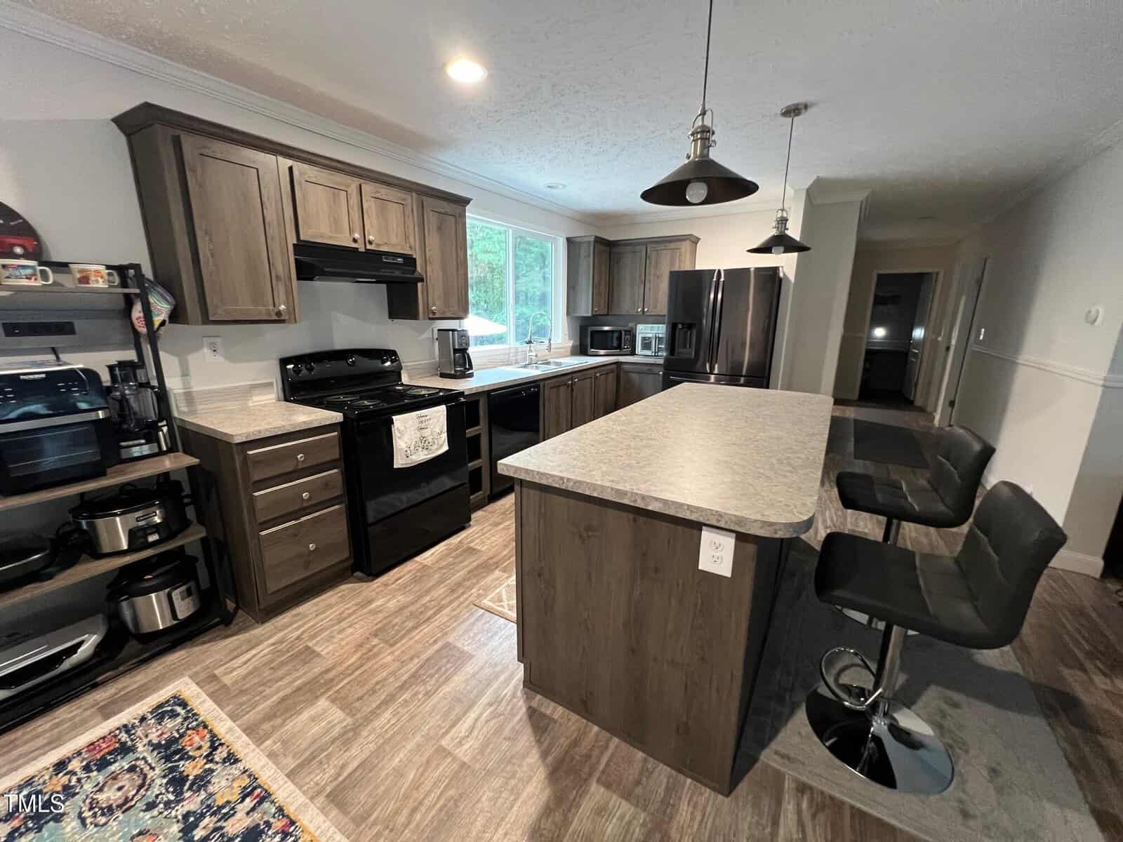 A modern kitchen with dark wood cabinets, black appliances, a large island with two black barstools, and various small appliances on the counters. There is a rug on the floor and recessed lighting overhead.