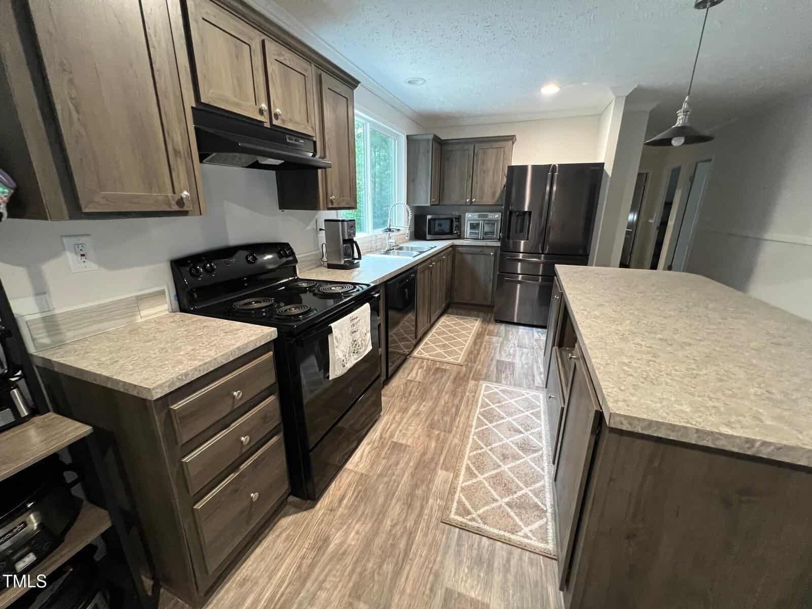 Modern kitchen with wood cabinets, black appliances, microwave, refrigerator, and a large island. Neutral countertops and two beige runners cover the wood-look flooring. Large window above the sink provides natural light.