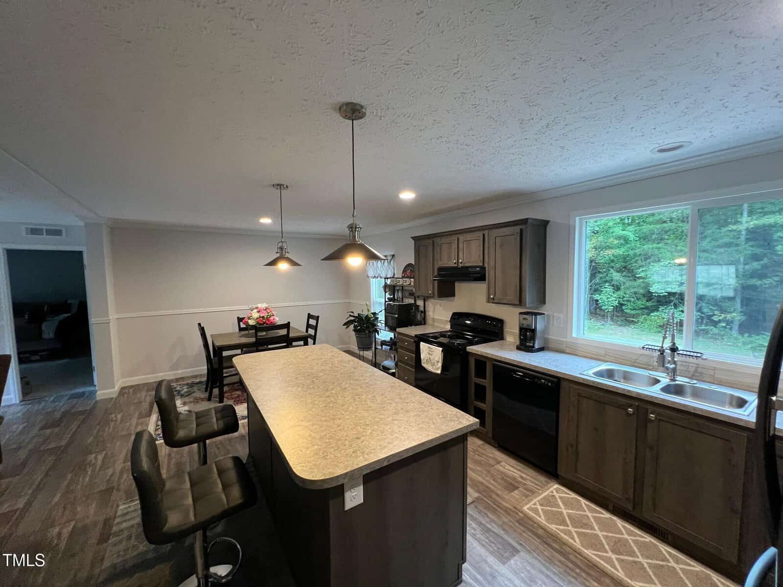 A modern kitchen with brown cabinets, black appliances, a center island with two bar stools, pendant lights, a dining table with flowers, and large windows showing greenery outside.
