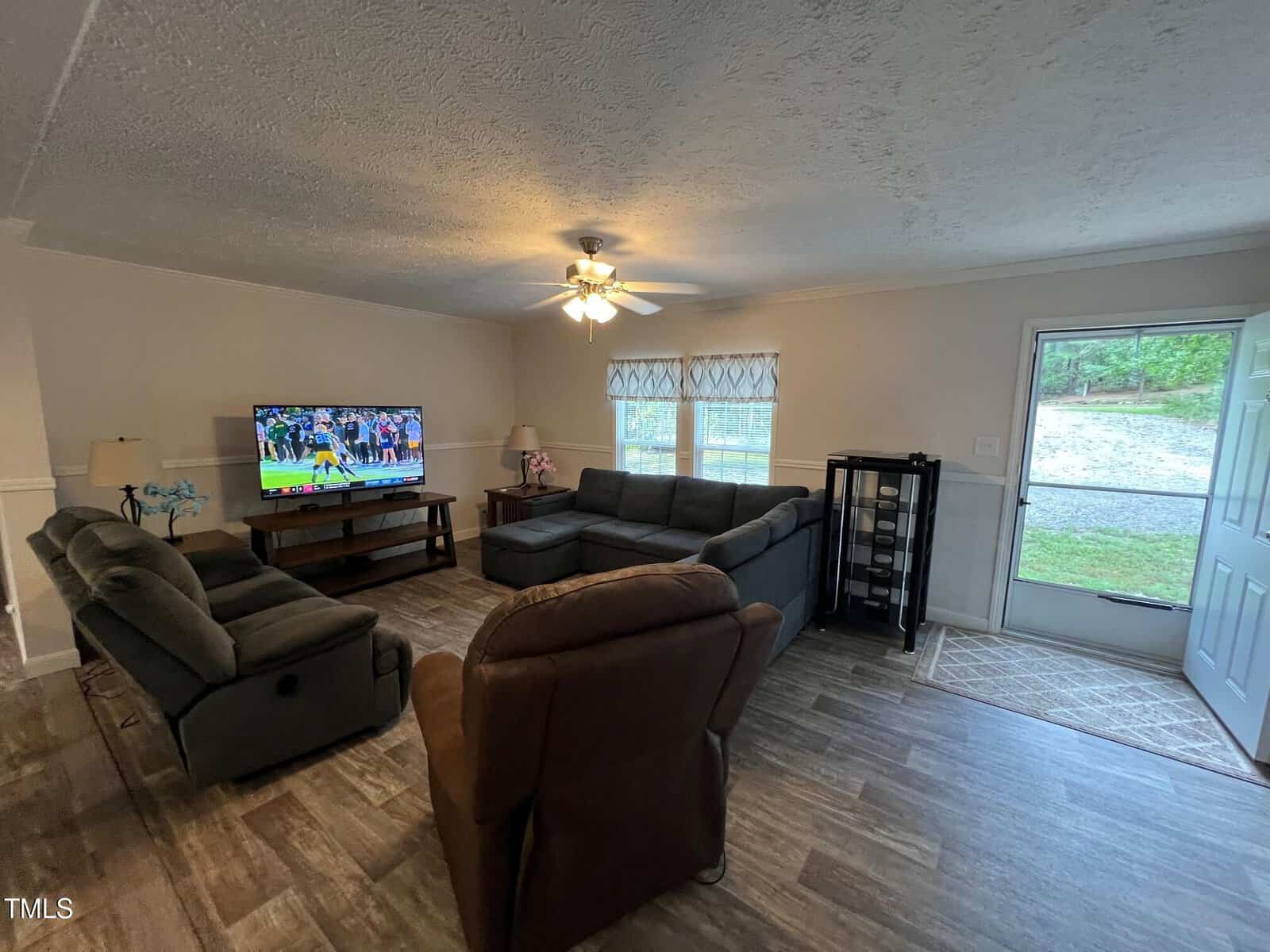 A living room with two brown recliners, a sectional sofa, a TV showing a football game, a coffee table, a ceiling fan with lights, and a door leading outside. The floor is wood and natural light enters through windows.