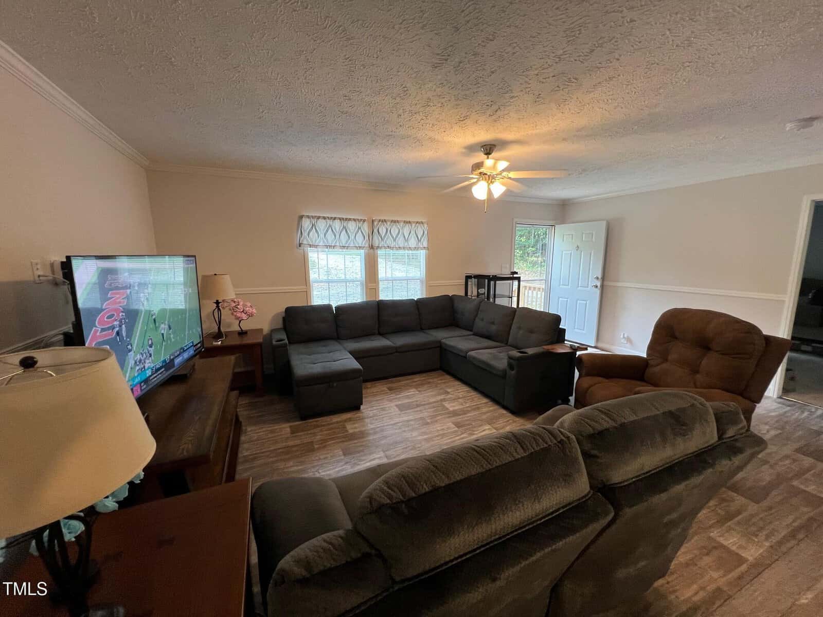 A living room with a sectional sofa, recliner, TV on a wooden stand, side tables with lamps, ceiling fan with light, two windows with curtains, and an open door showing a porch outside.
