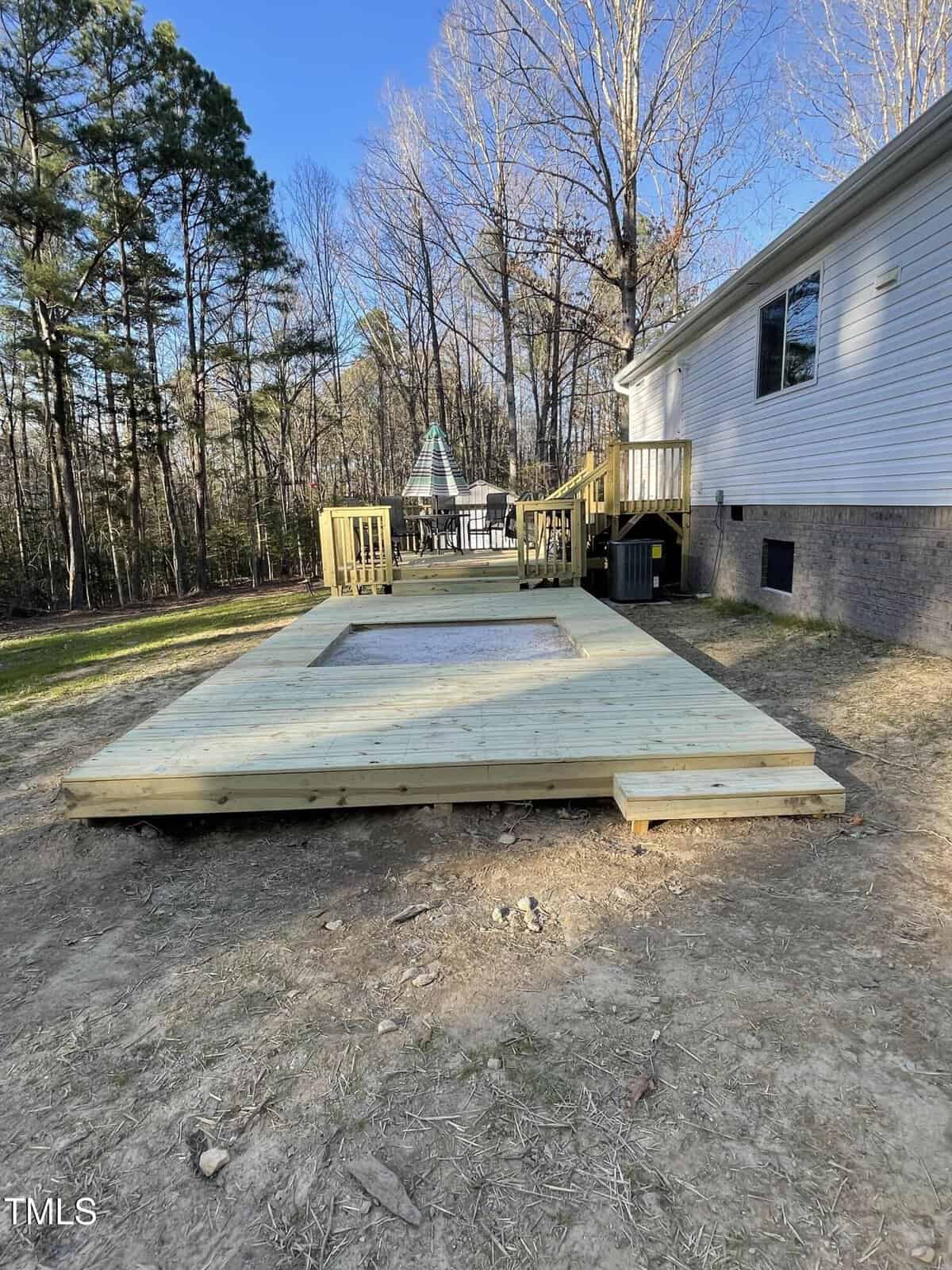 A raised wooden deck extends from a house into a backyard, featuring a rectangular recessed area in the center. Stairs lead from the house to the deck, surrounded by trees and bare ground.