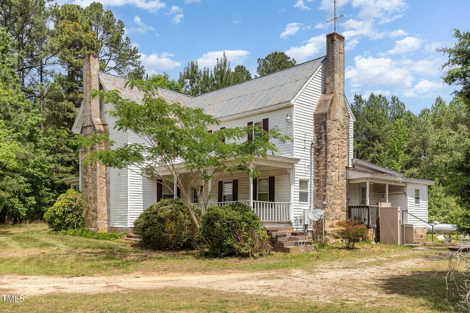 Nestled on 5062 Dorsey Road in Oxford, this two-story white farmhouse features a covered front porch, exposed stone chimneys on each end, a metal roof, and is surrounded by trees and greenery under a blue sky with scattered clouds.