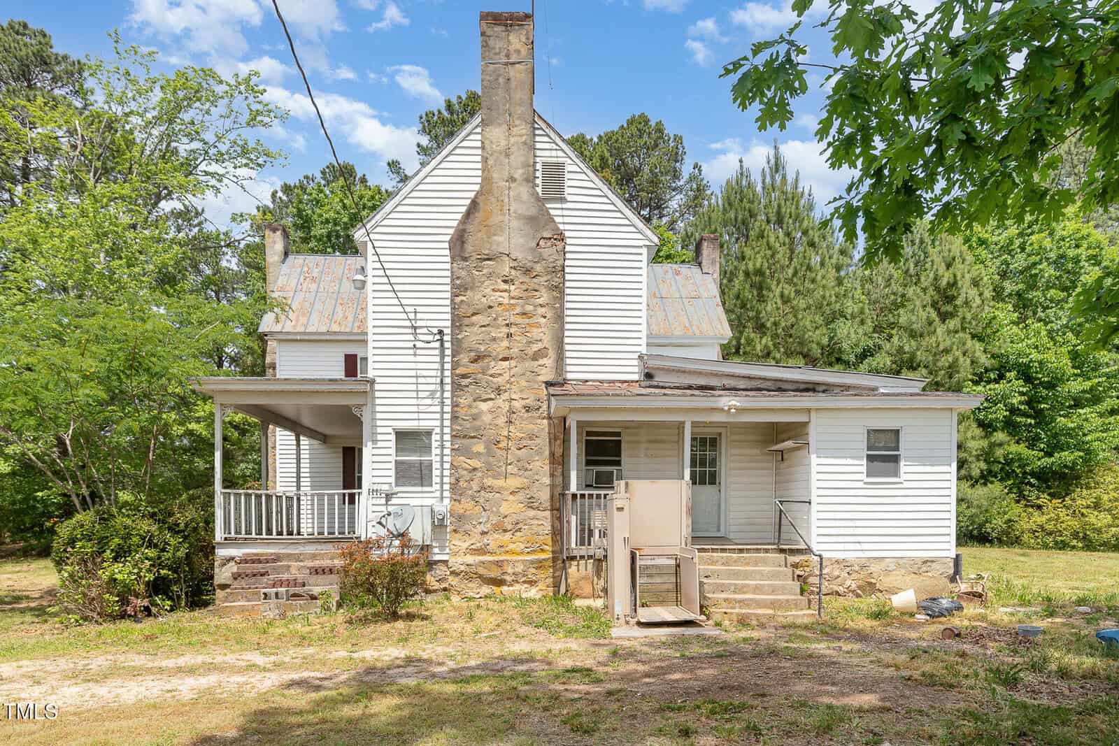 A white two-story house with a large stone chimney, a covered porch, and an exterior staircase leading to a back door. The yard is grassy with scattered trees, and there are some items beside the steps.