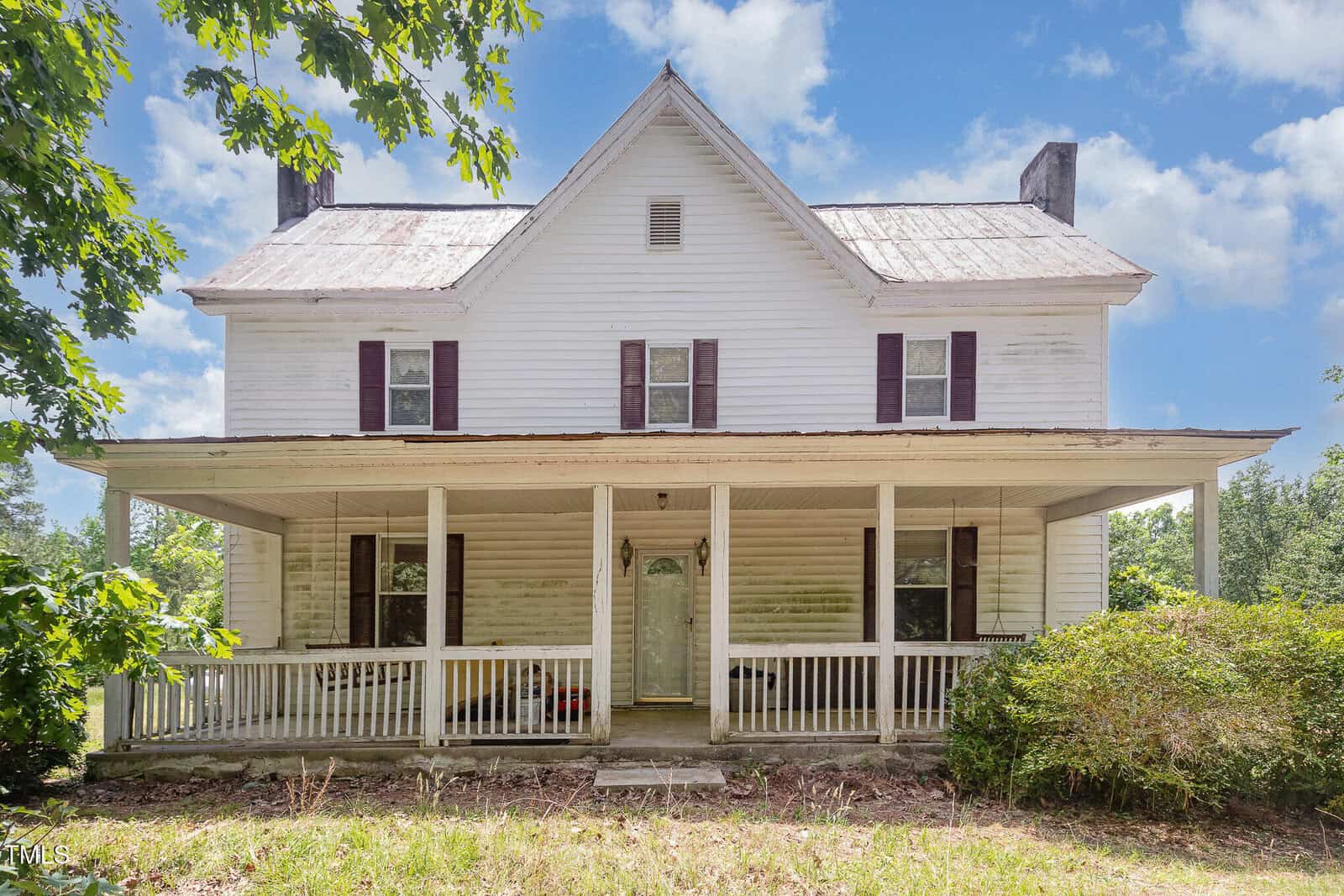 A two-story white house with a metal roof, two chimneys, and a wide front porch. The house has two upper windows with brown shutters and visible weathering. Bushes and trees are in the yard, under a blue sky with clouds.