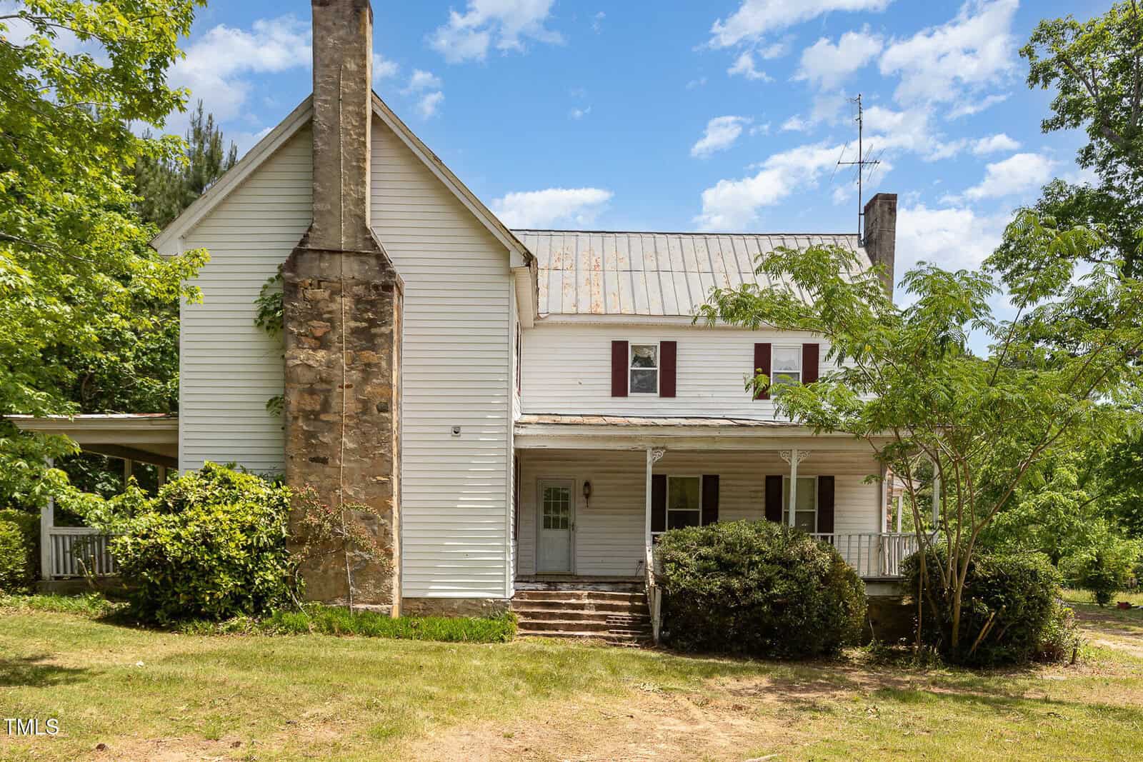 A two-story white house with a metal roof, large stone chimney, covered front porch, and wooden steps, surrounded by trees and bushes under a partly cloudy sky.
