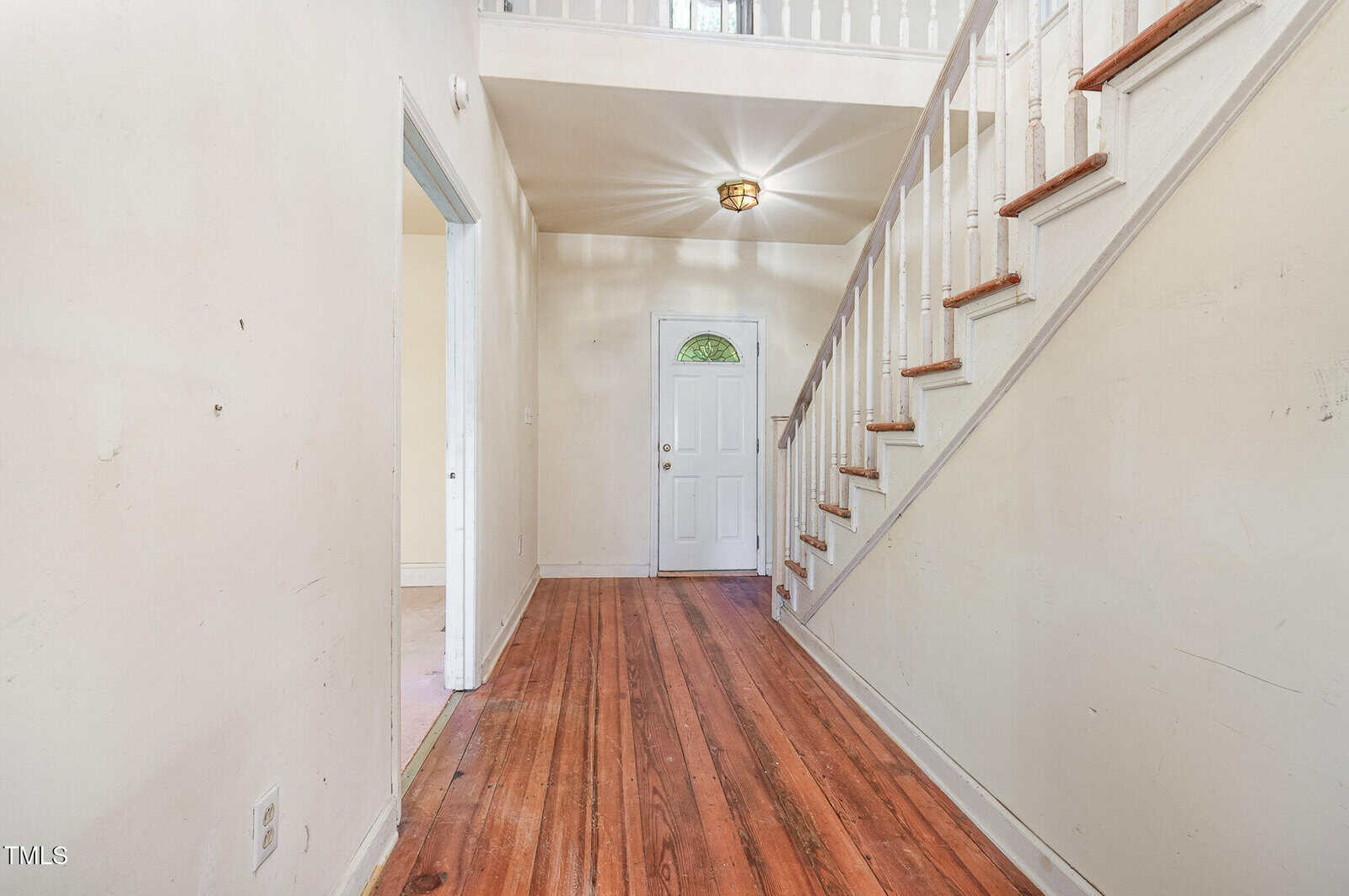 A hallway with hardwood floors, cream-colored walls, a white staircase with wooden handrails, and a white front door with a small arched window. A light fixture is on the ceiling above the entry.