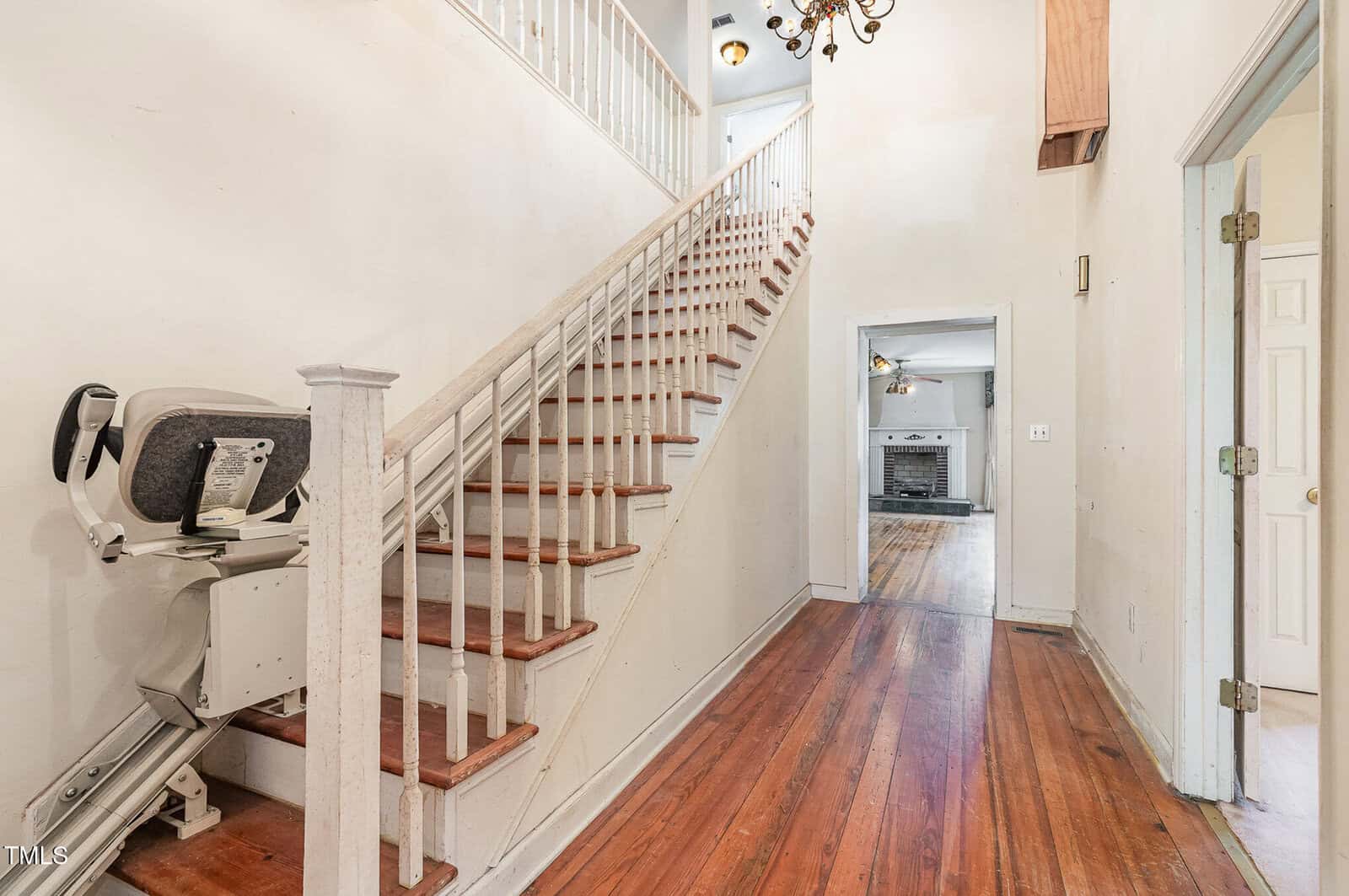 A hallway with wooden floors and a staircase with a stair lift on the left; a room with a fireplace is visible straight ahead, and partially open doors are on the right side of the hallway.