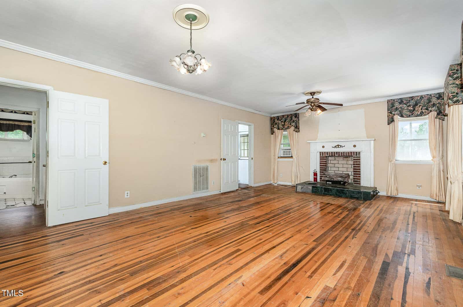Spacious living room with hardwood floors, beige walls, a white fireplace with green marble hearth, ceiling fan, chandelier, and large windows with patterned valances and curtains. Doorways lead to adjoining rooms.