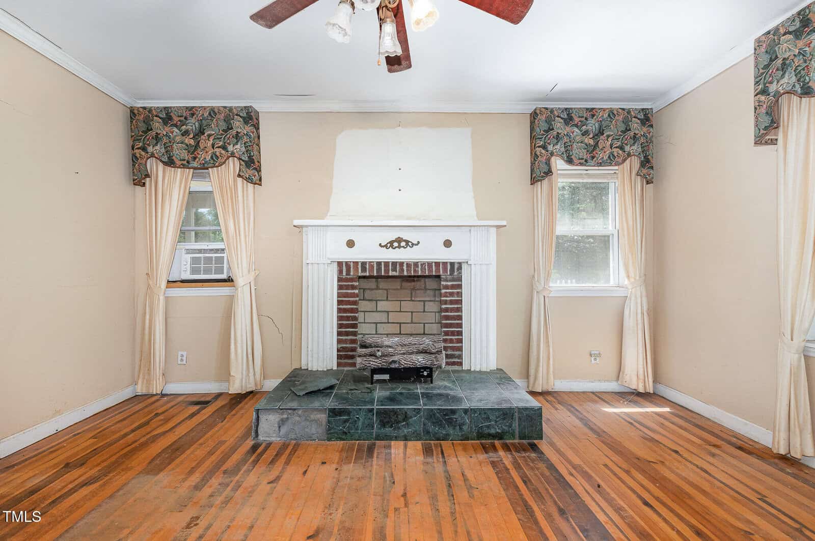 A living room with hardwood floors, a white fireplace with a green marble hearth, two windows with floral valances and beige curtains, and a ceiling fan with light. The wall above the fireplace is unfinished.