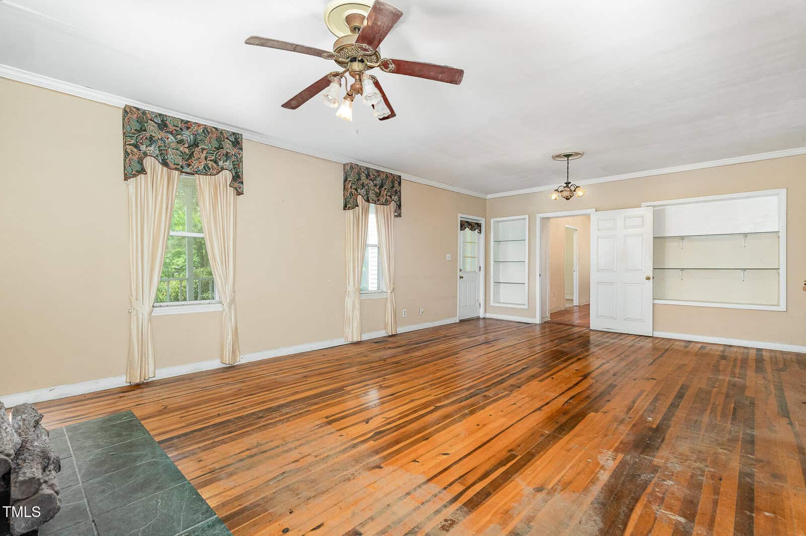 A spacious living room with hardwood floors, beige walls, two windows with cream curtains and valances, ceiling fans with lights, built-in shelves, and a green-tiled corner area. The room appears empty.