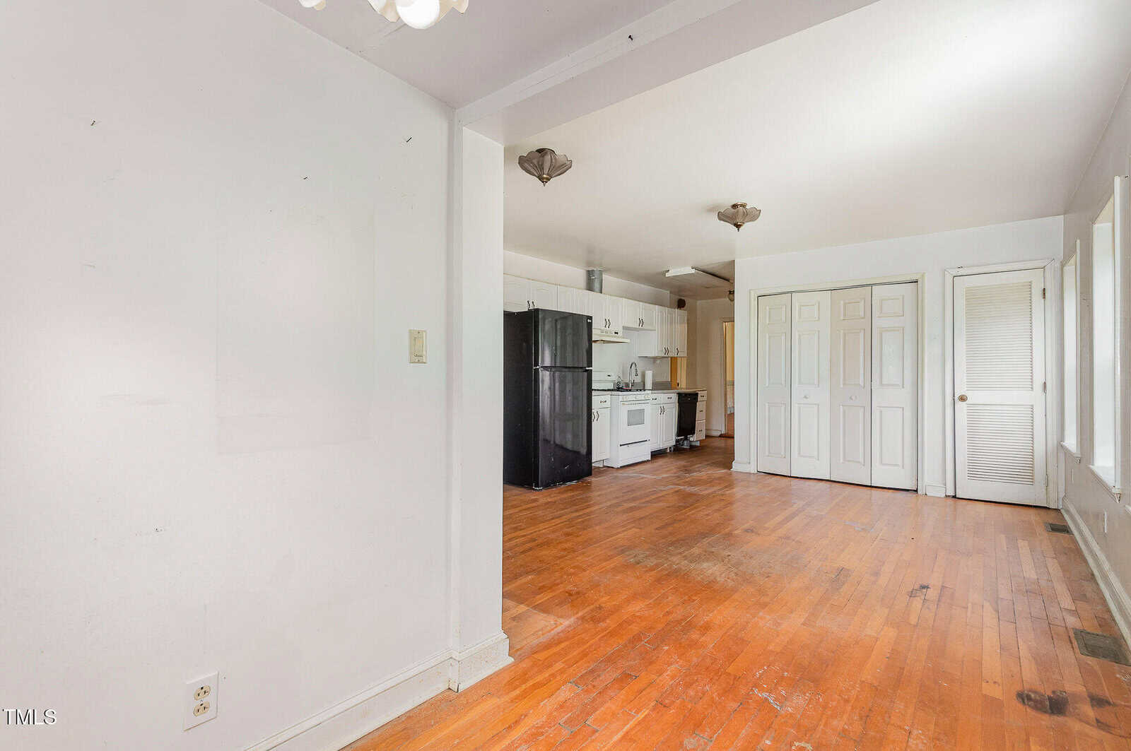 A mostly empty room with hardwood floors, white walls, a small kitchen area with white cabinets and black appliances, double closet doors, and several windows letting in natural light.