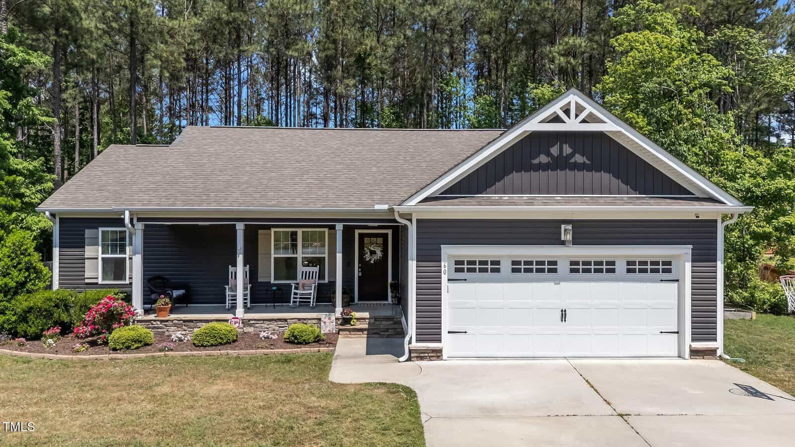 Single-story house with dark gray siding, white trim, and a two-car garage. A small front porch with white railings and a bench sits amid landscaped bushes, with tall trees in the background and a concrete driveway in front.