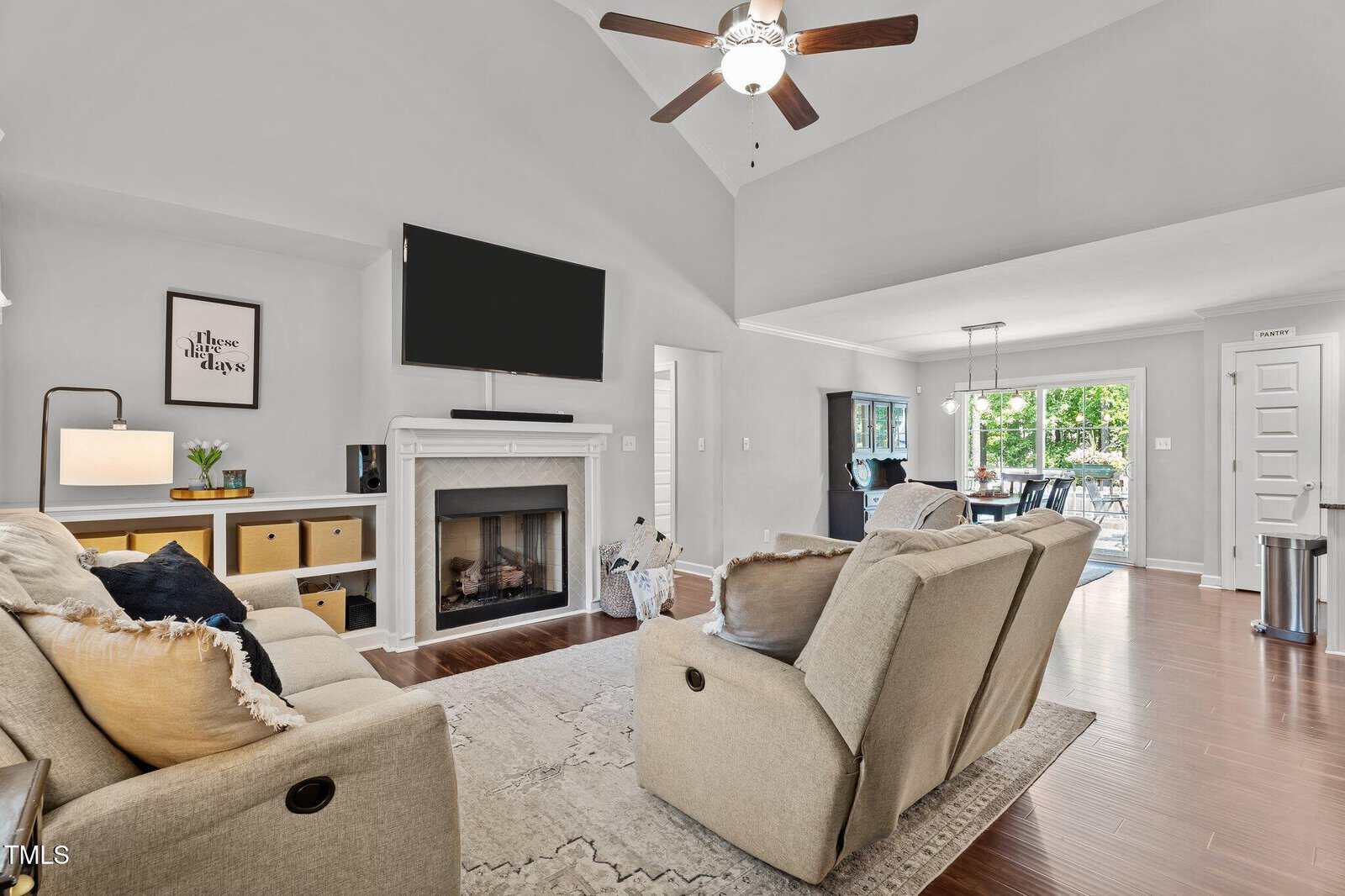 A modern living room features a beige sofa and two recliners facing a wall-mounted TV above a fireplace, with wooden floors, a cozy rug, and an open view to the dining area and backyard.
