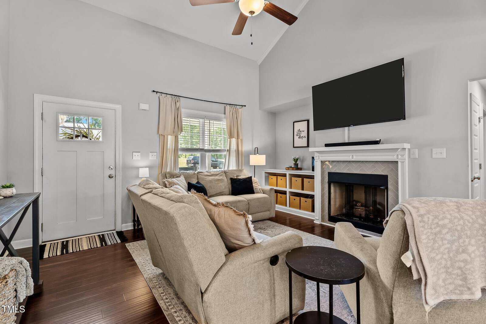 A cozy living room with beige sofas, a large wall-mounted TV above a fireplace, a ceiling fan, hardwood floors, and a window letting in natural light. Shelves and plants accent the space.