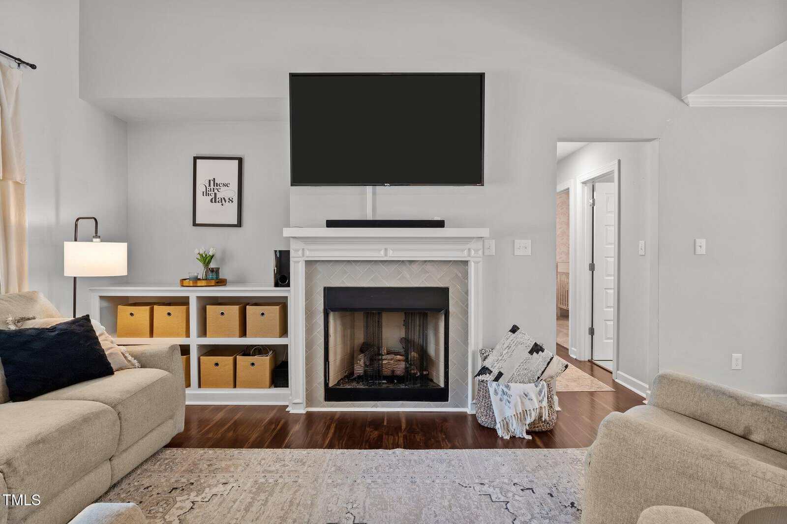 A cozy living room with a fireplace beneath a mounted TV, flanked by storage cubes with baskets, beige sofas, a framed quote on the wall, a lamp, and a doorway leading to another room.