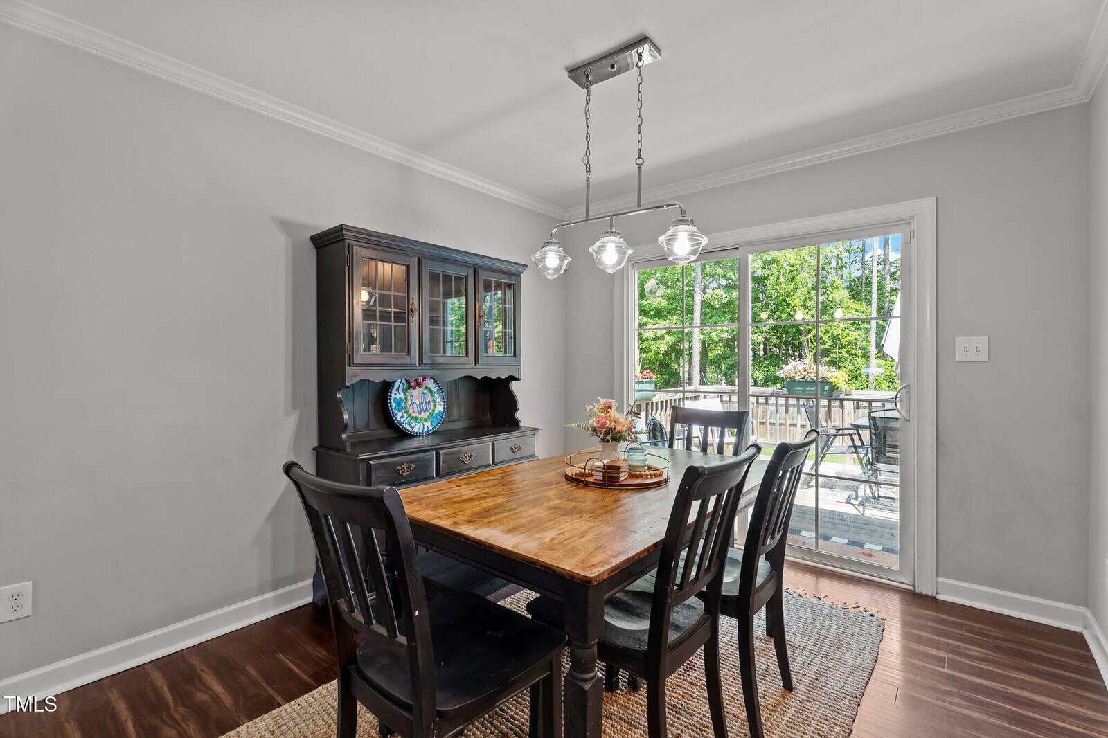 Dining room with a wooden table, four black chairs, a black hutch, and a woven rug. A light fixture hangs above the table. Glass doors open to a deck with outdoor furniture and green trees visible outside.
