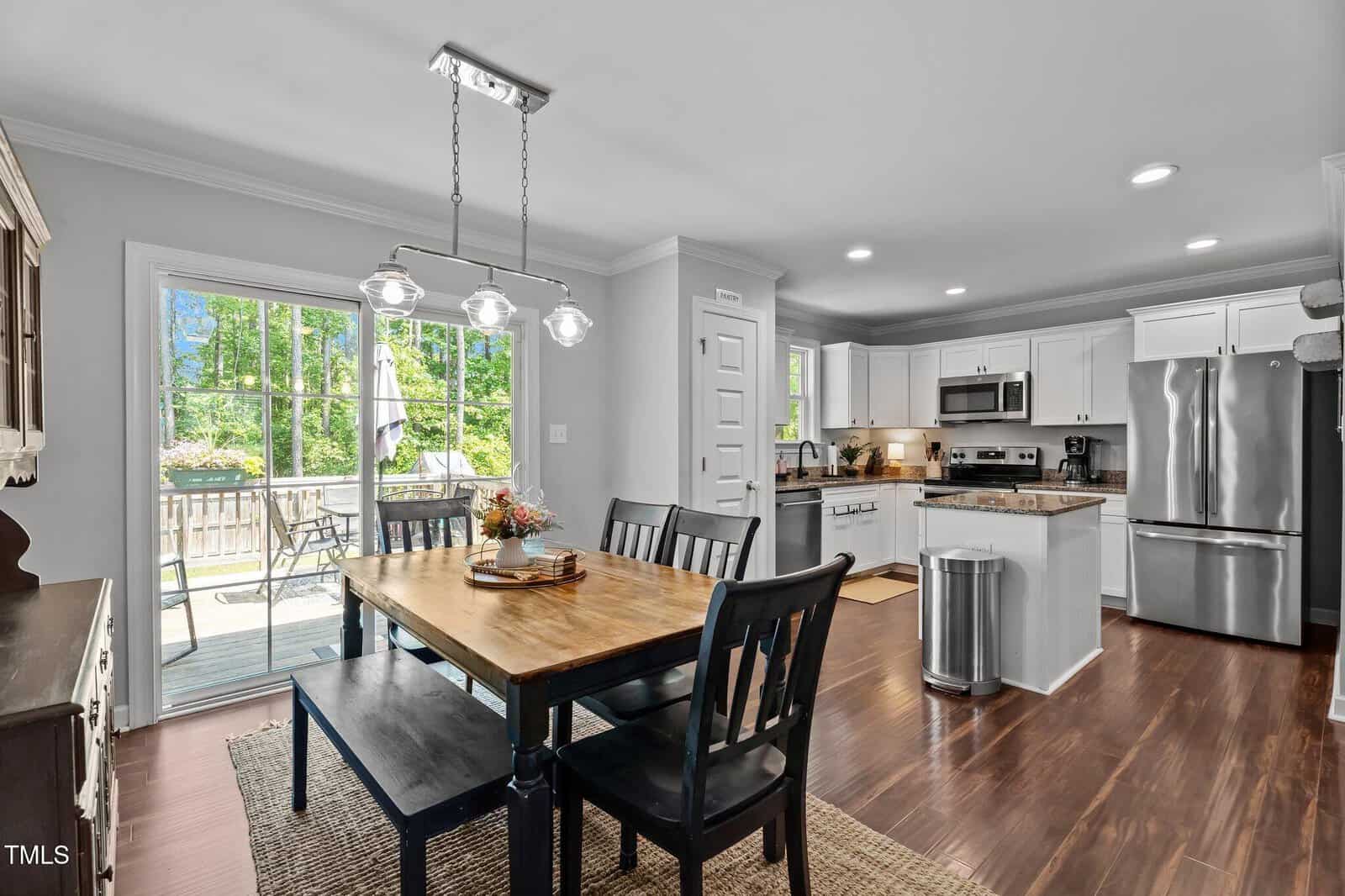 A modern kitchen and dining area with white cabinets, stainless steel appliances, a wooden dining table with black chairs and bench, hanging lights, and a sliding glass door leading to a deck with outdoor seating.