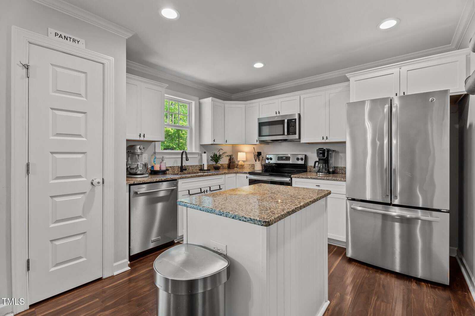 Modern kitchen with white cabinets, stainless steel appliances, granite countertops, a small island with a trash can, hardwood floor, and a door labeled “Pantry.” A window above the sink lets in natural light.