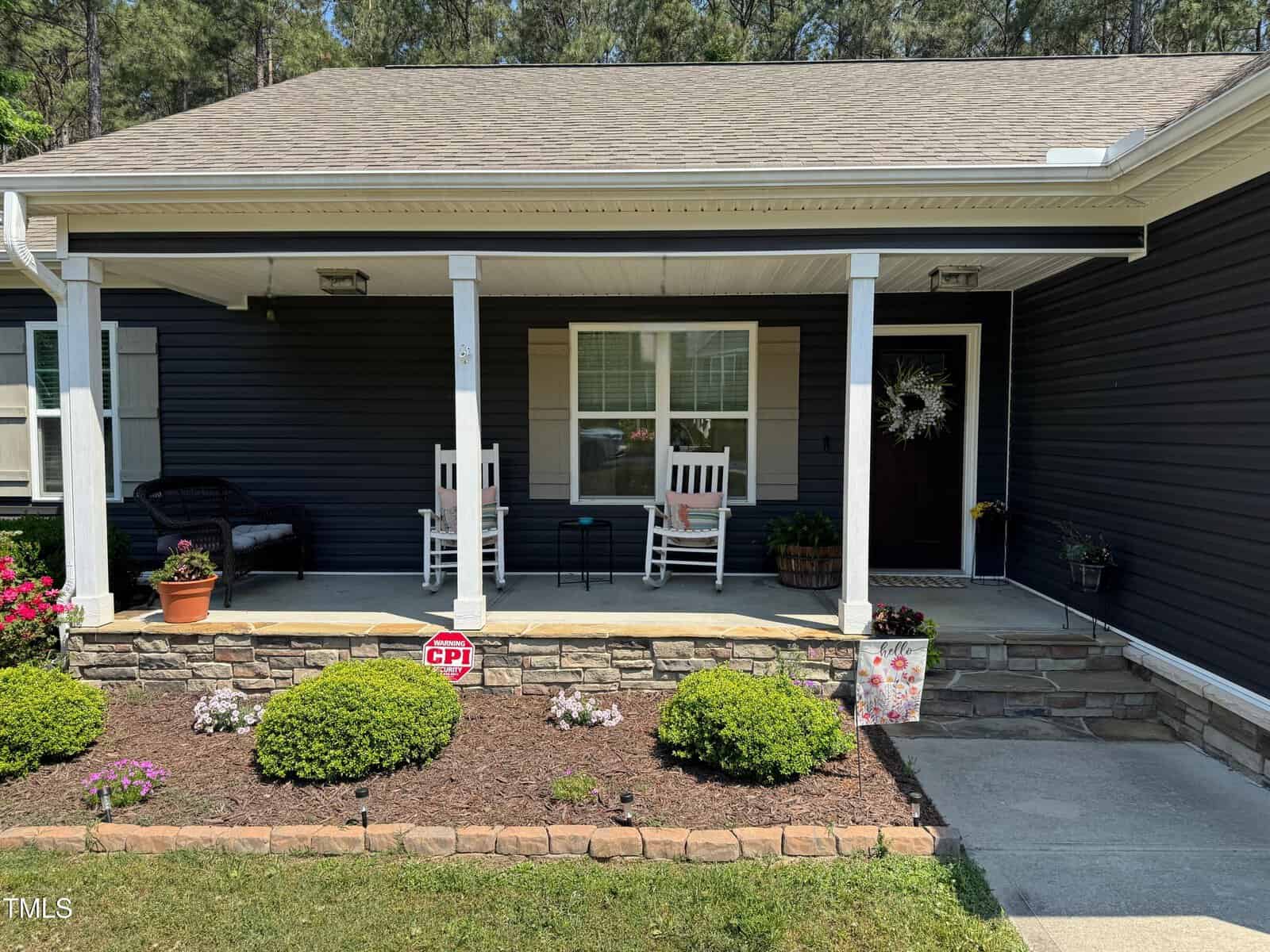 A house front porch with dark siding, white trim, two white rocking chairs, a small table, a bench, potted plants, a wreath on the door, a CPI security sign, and neatly trimmed bushes in the garden.