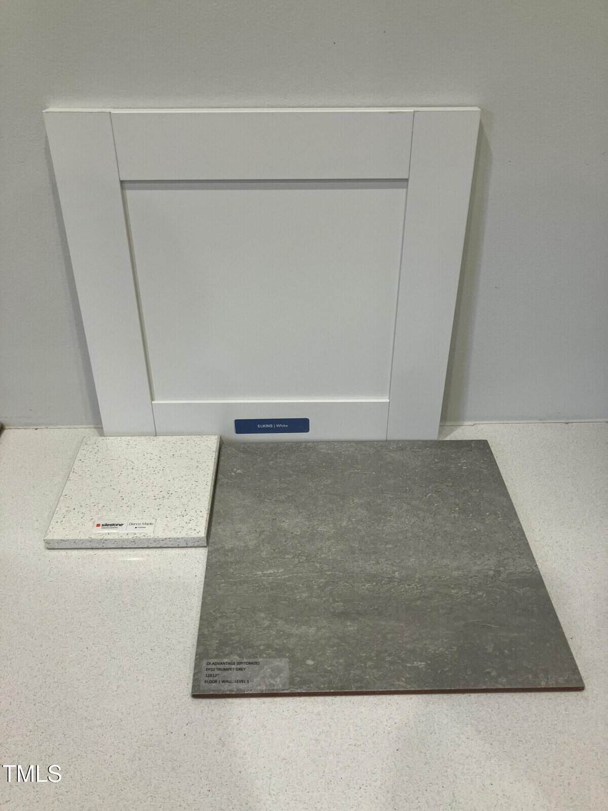 A white cabinet door stands upright against a wall, with a square white countertop sample and a larger square gray floor tile sample placed in front of it on a white surface.