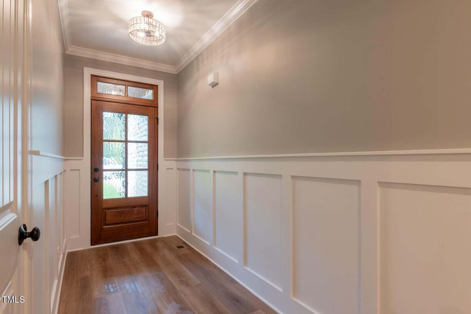 A hallway with light wood floors, white wainscoting, light gray upper walls, and a wooden front door with frosted glass panels. A modern chandelier hangs from the ceiling, lighting the space.
