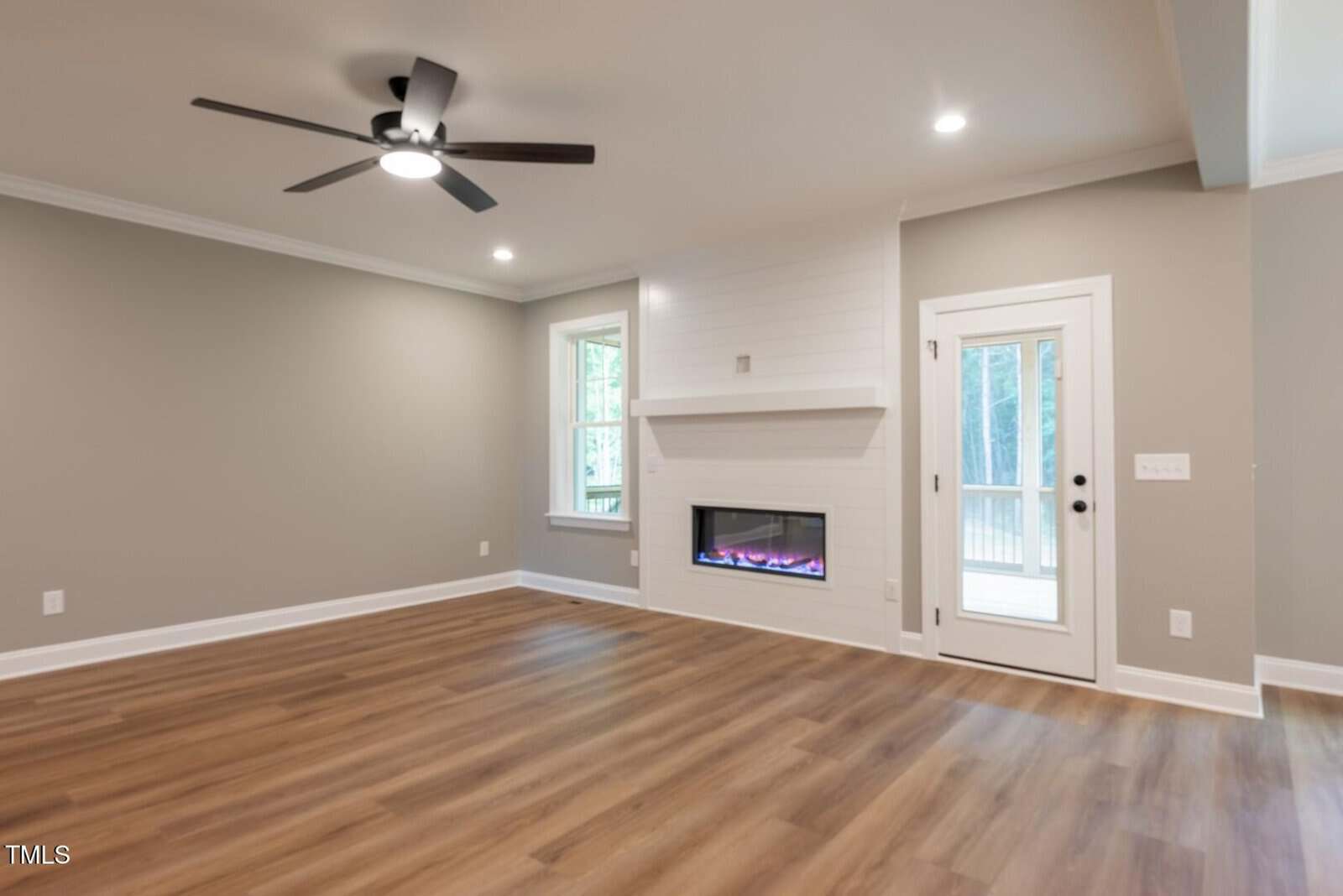 A modern, empty living room with light wood flooring, gray walls, white trim, a ceiling fan, a window, a glass door, and a built-in electric fireplace with a white mantel.