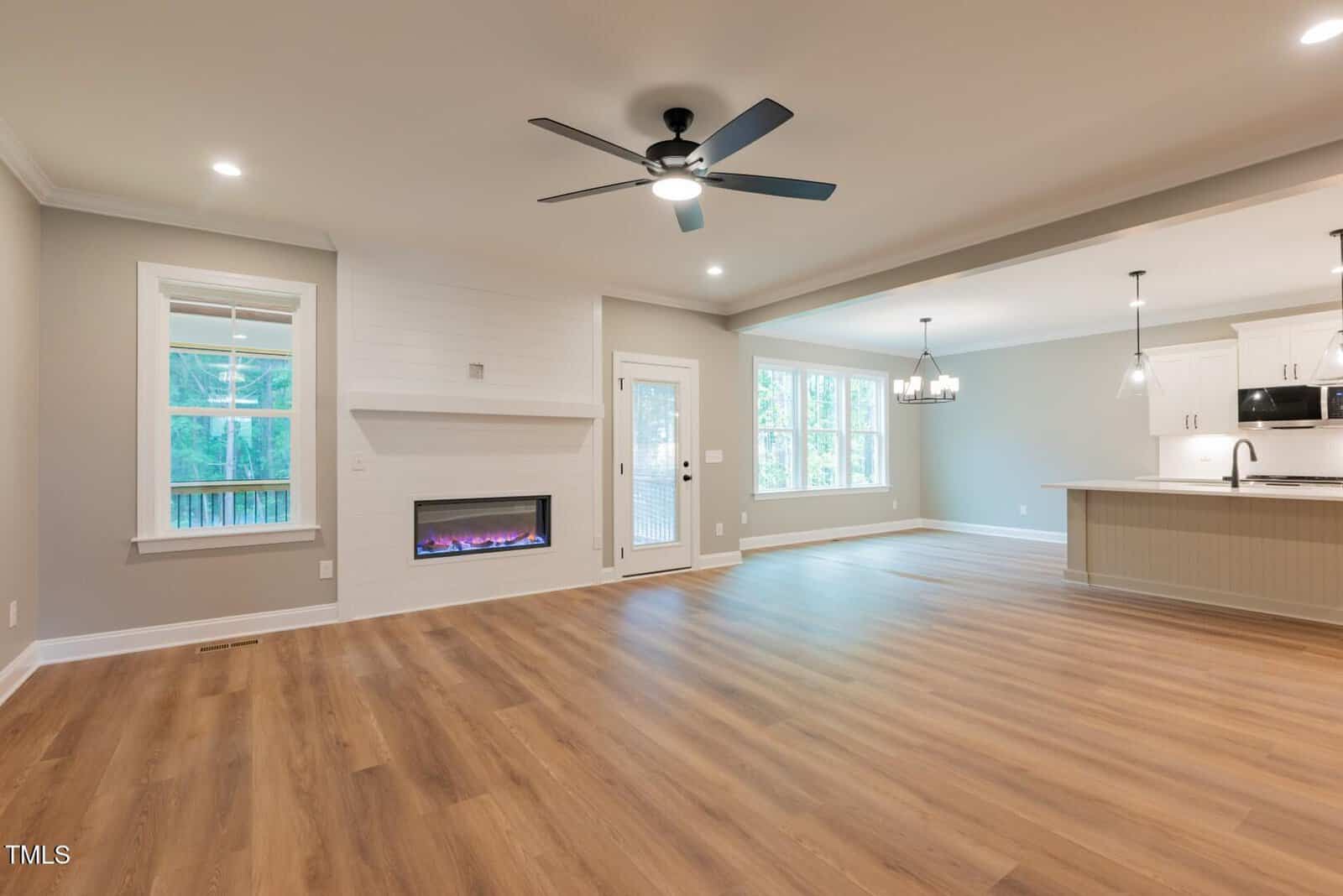 Spacious, modern living and dining area with wood floors, ceiling fan, electric fireplace, large windows, and open kitchen with white cabinets, black fixtures, pendant lighting, and recessed lights.
