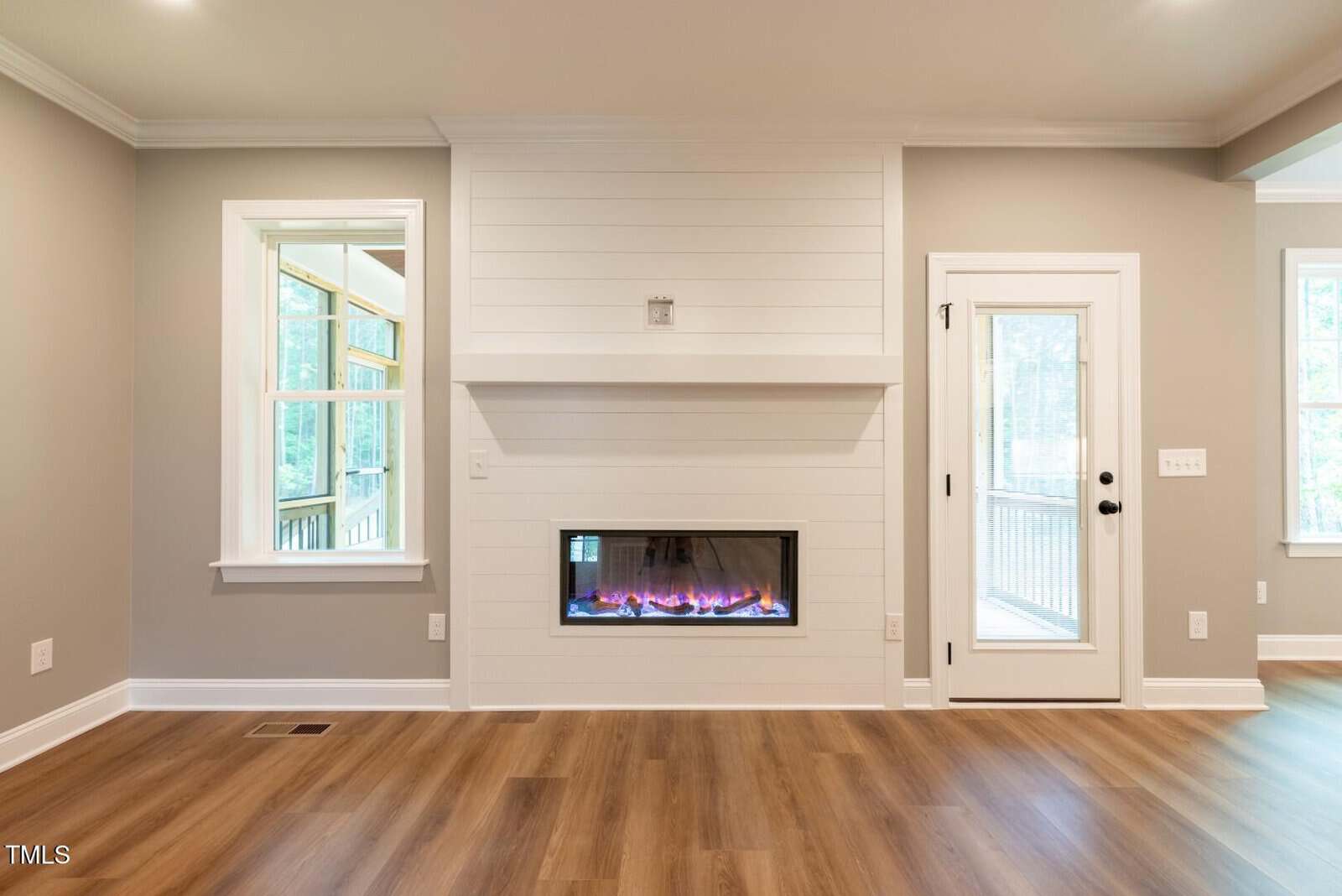 Modern living room with light wood floors, gray walls, a horizontal electric fireplace with shiplap paneling, large windows on the left, and a glass door on the right leading outside. Room is unfurnished.