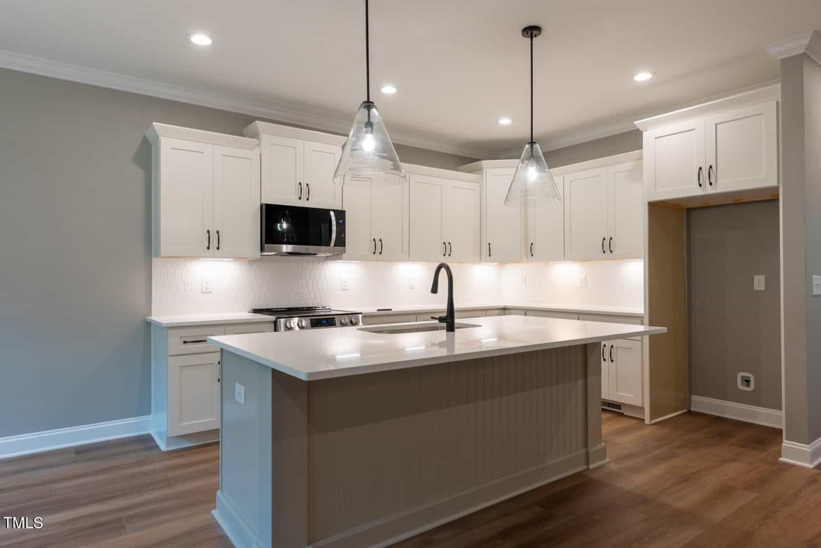 Modern kitchen with white cabinets, stainless steel appliances, center island with sink, pendant lights, light wood floor, and recessed ceiling lights. An empty alcove is visible on the right wall.