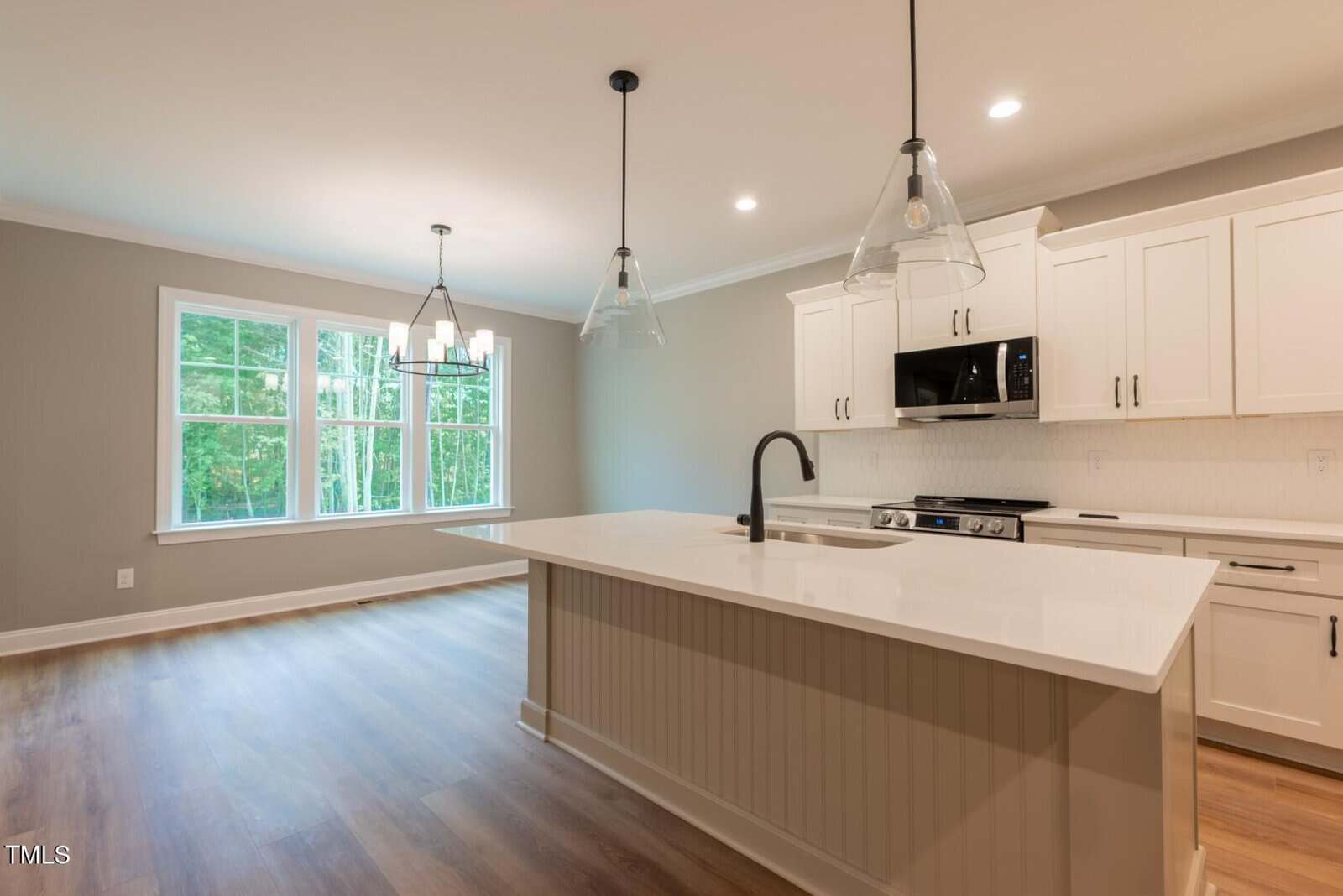 Modern kitchen with white cabinets, an island with sink, stainless steel appliances, pendant lights, and adjacent dining area with large windows and wood flooring. Walls are painted light gray.