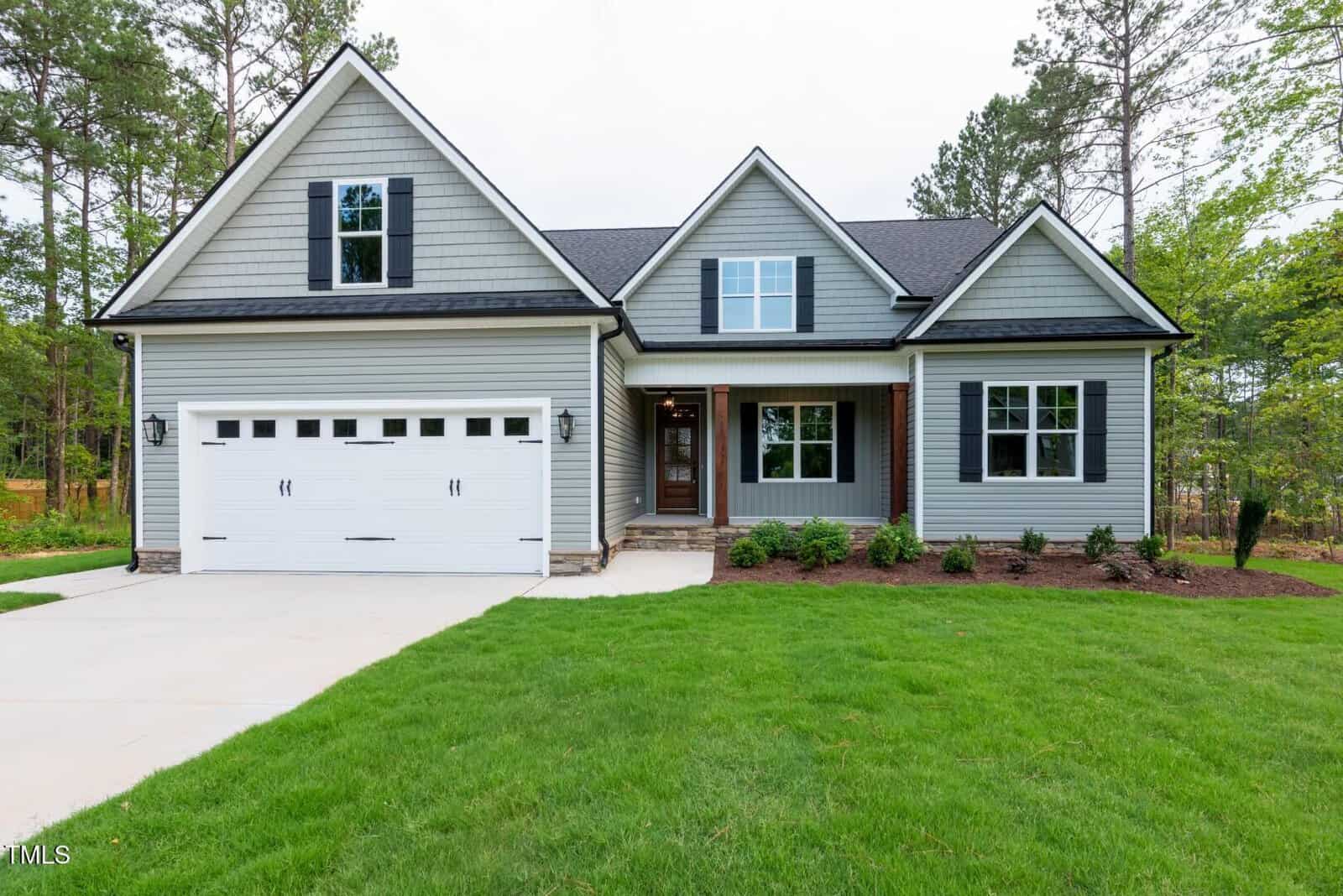 A modern suburban house with gray siding, black shutters, a two-car garage, and a manicured lawn, surrounded by trees and greenery on a cloudy day.