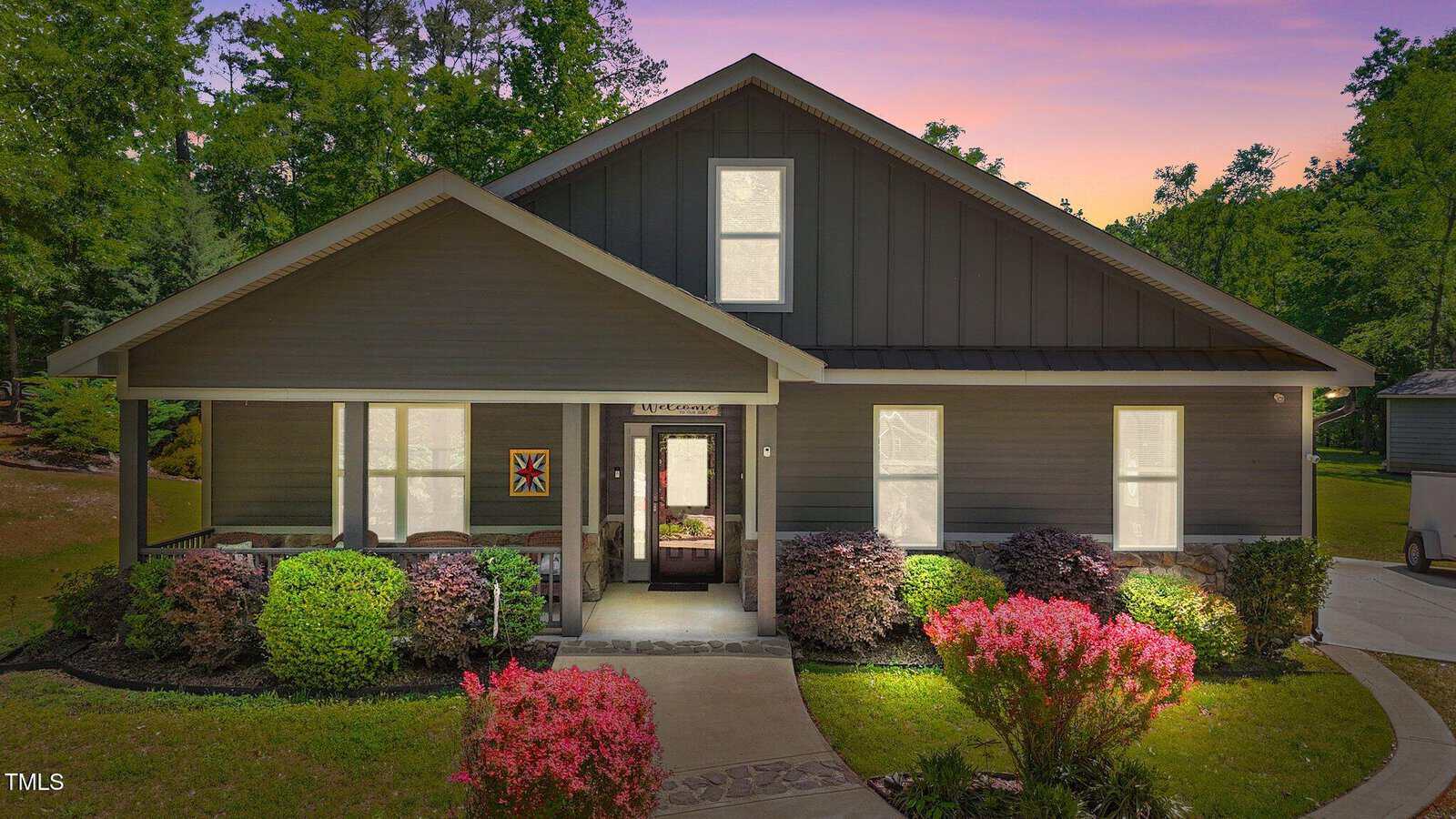 A modern single-story house with dark gray siding, white trim, a covered front porch, and large front windows, surrounded by neatly trimmed bushes and pink flowering shrubs at sunset.