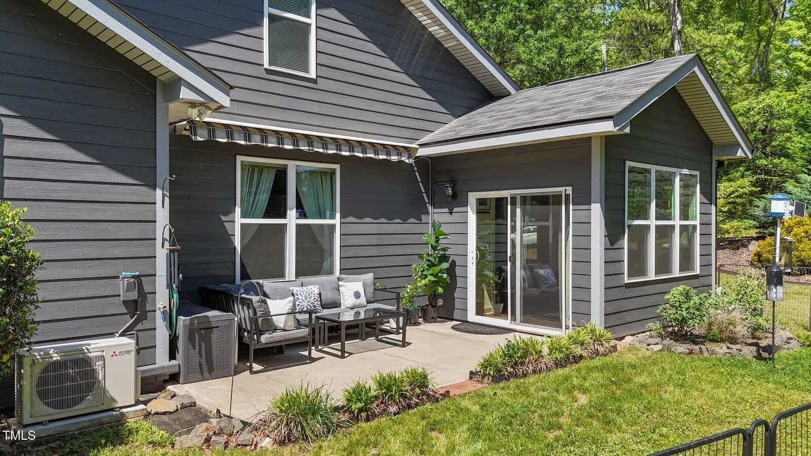 A gray house with white trim has a small concrete patio featuring outdoor seating, a table, potted plant, sliding glass door, windows, awning, and central AC unit, bordered by grass and landscaping. Trees are visible in the background.