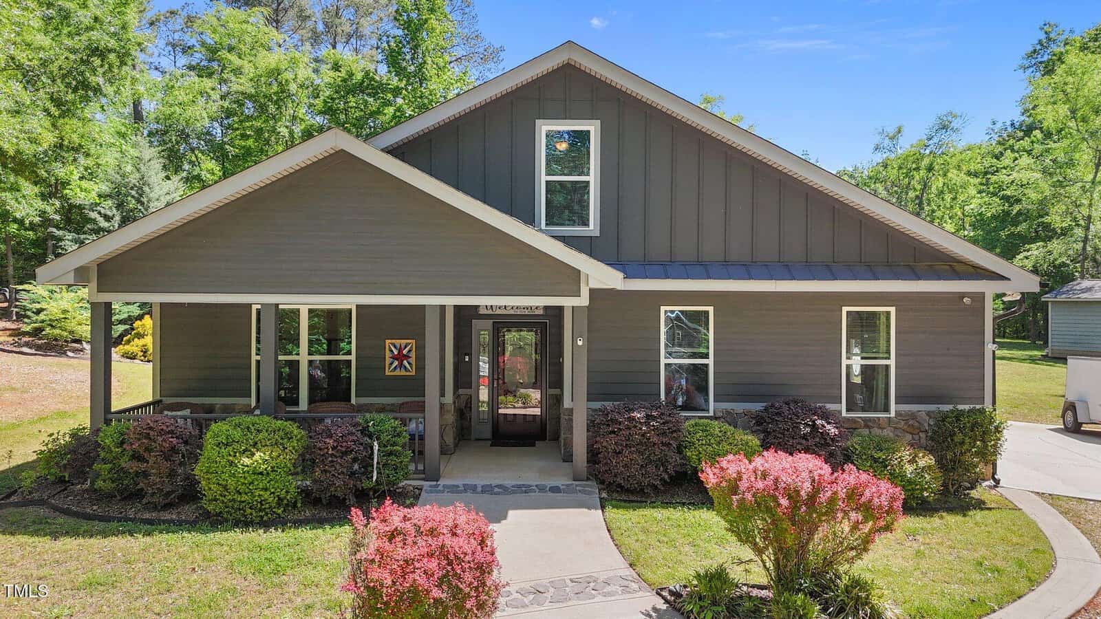 A modern gray house with a large front porch, white trim, and neatly trimmed bushes in the front yard on a sunny day, surrounded by trees.