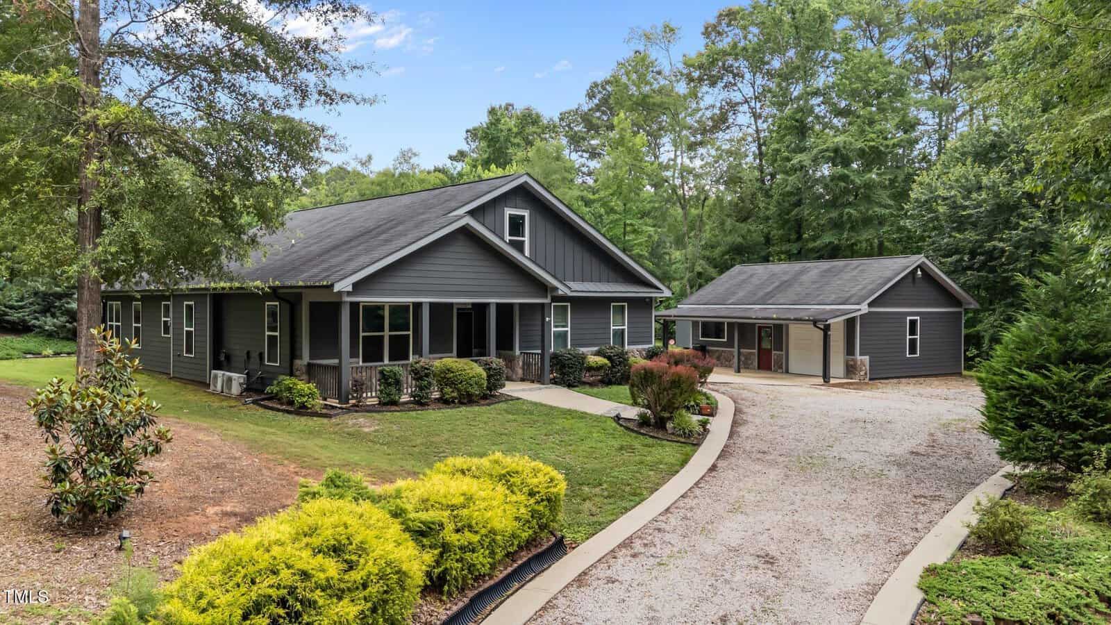 A modern dark gray house with a gabled roof, front porch, and stone accents sits beside a detached garage, surrounded by trees, landscaped bushes, and a gravel driveway on a green lawn under a partly cloudy sky.