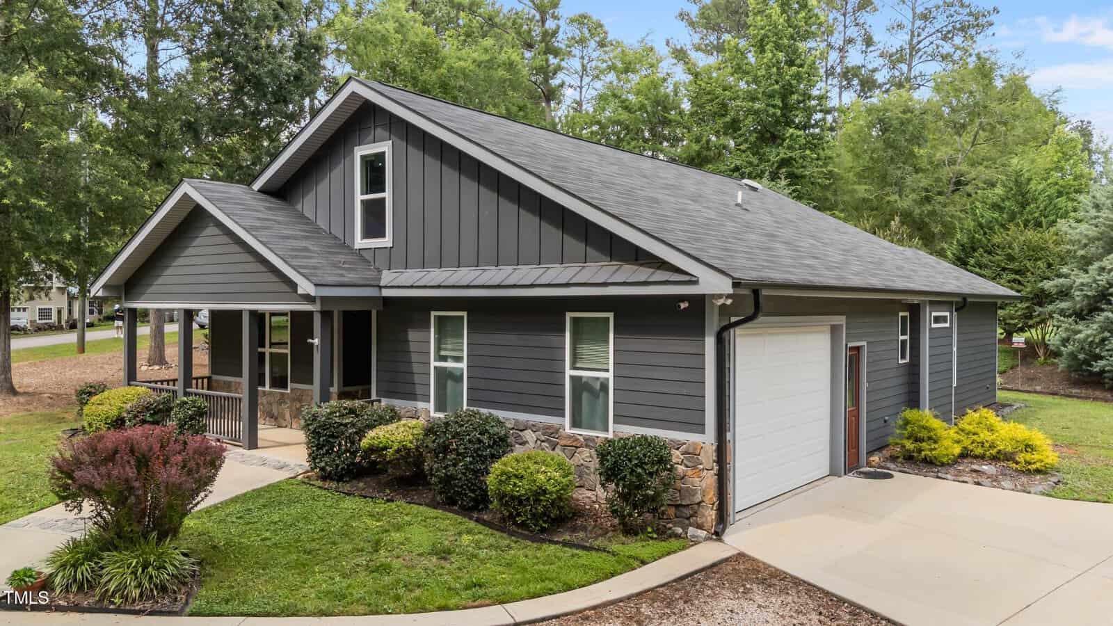 A modern, single-story house with dark gray siding, white trim, and a small front porch. The home features a one-car garage and is surrounded by greenery, shrubs, and mature trees.