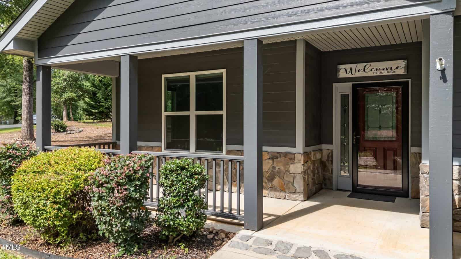 A covered porch with gray siding, stone accents, a window, and a glass front door with a "Welcome" sign above it. There are green shrubs and a small railing along the edge of the porch.