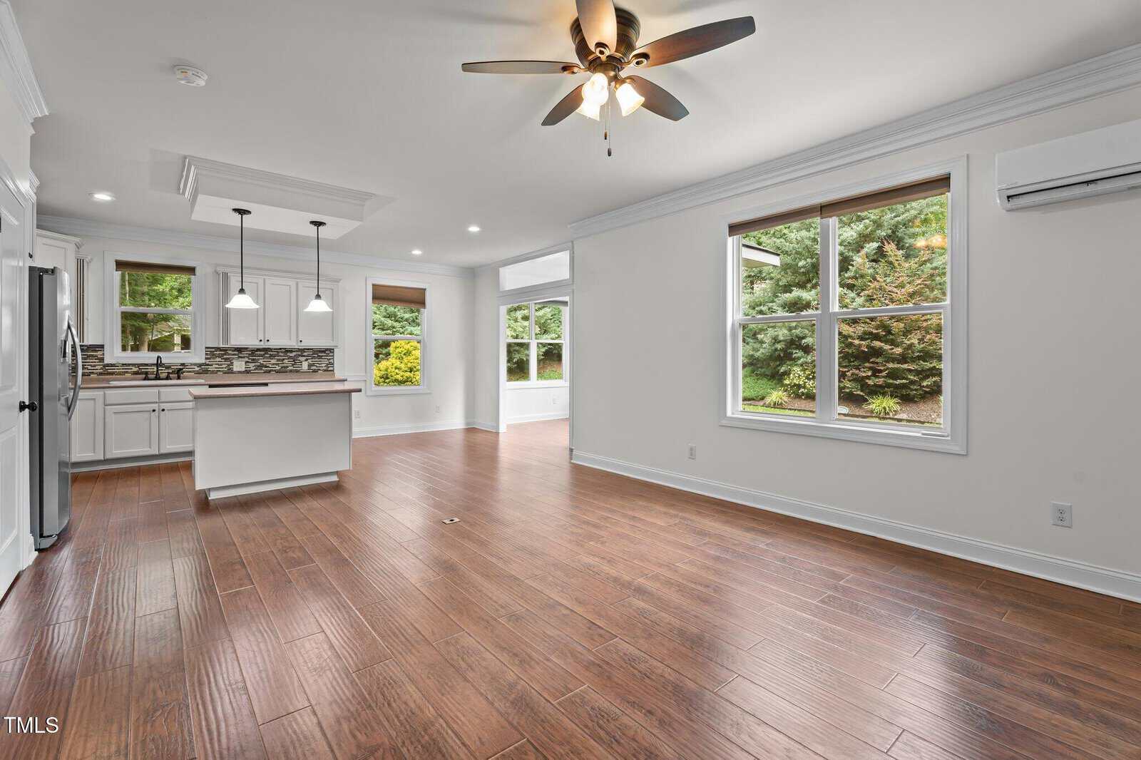 Open living area with wood floors, white walls, ceiling fan, and large windows. Kitchen with white cabinets, stainless steel appliances, tile backsplash, and an island is visible in the background. Trees seen through the windows.