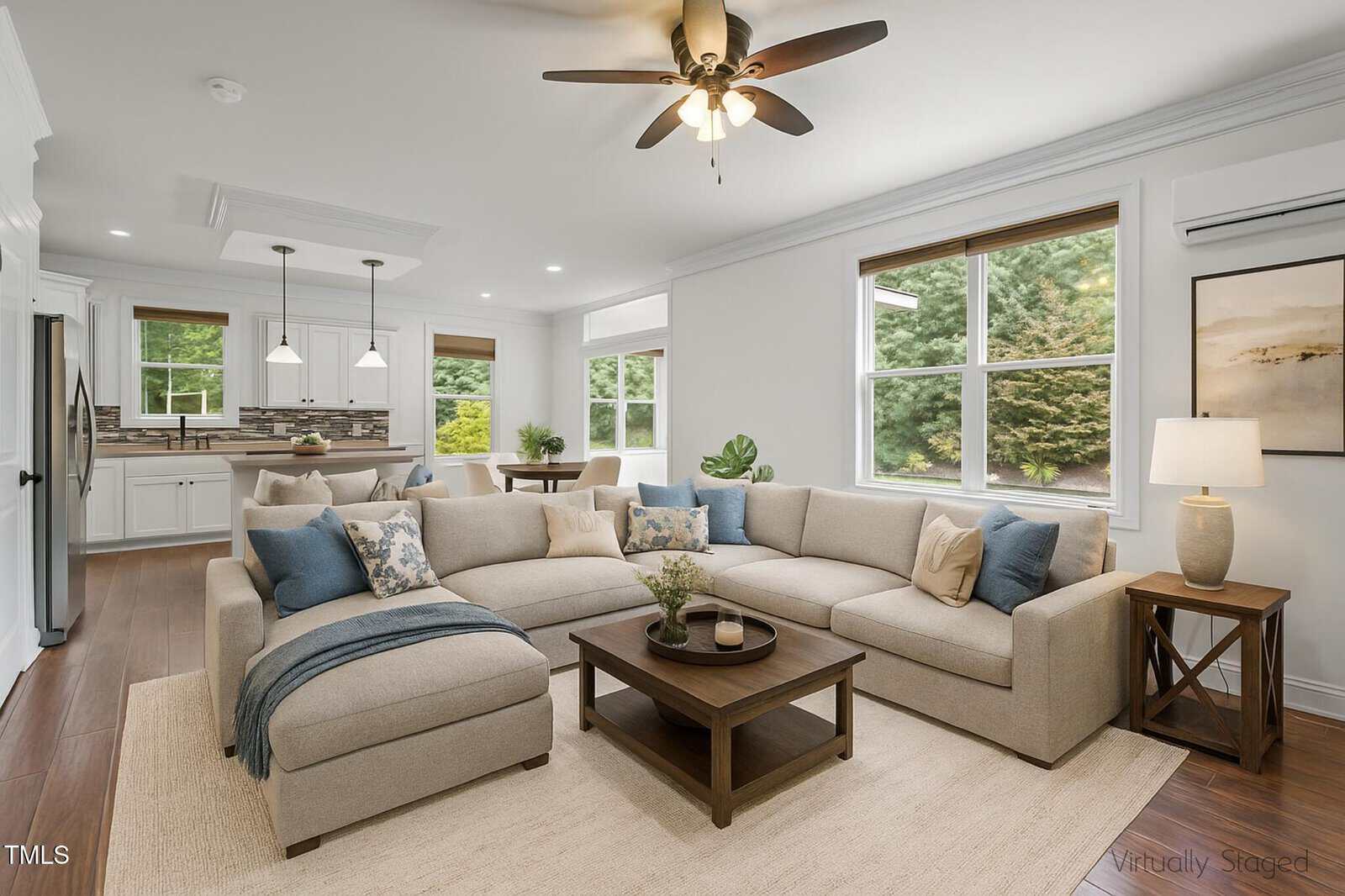 A bright living room with a beige sectional sofa, wooden coffee table, side table with a lamp, indoor plants, and large windows overlooking greenery; the open kitchen and dining area are visible in the background.