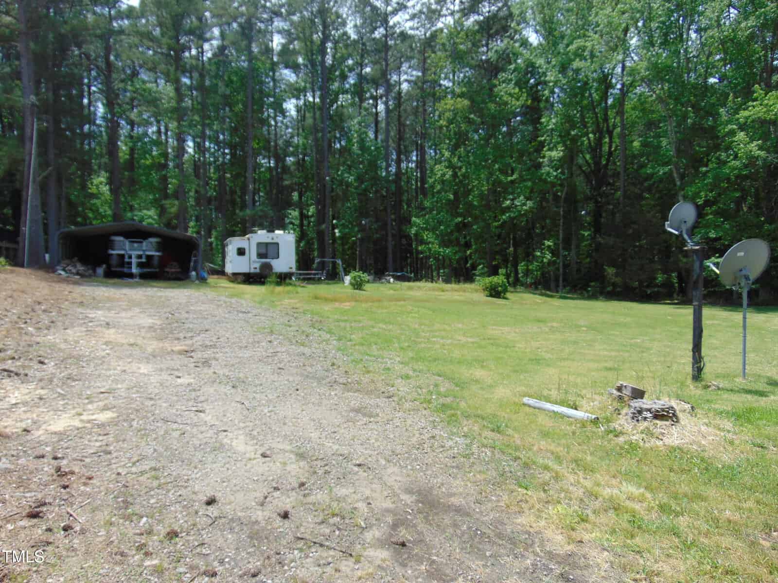 A gravel driveway leads to a small trailer and covered outdoor seating at 198 S Piney Grove Road, Manson. Set in a grassy clearing surrounded by trees, the yard also features two satellite dishes mounted on wooden posts.