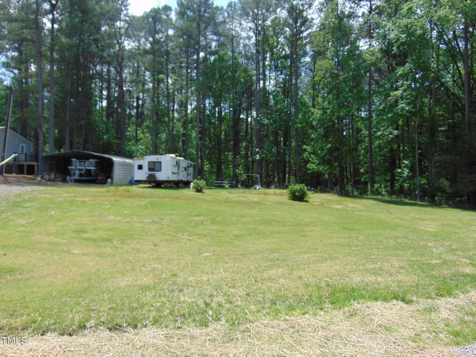 A grassy yard with a few small bushes, a white camper, a metal carport, and dense pine trees in the background. A gray house is partially visible on the left edge of the image.