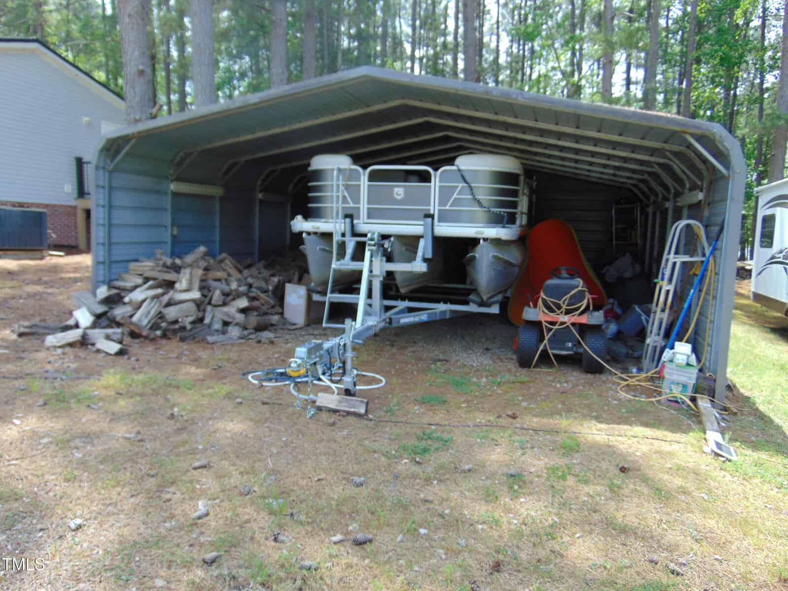 A covered metal carport shelters a pontoon boat, a stack of firewood, an orange kayak, a ladder, and various outdoor equipment. Tall trees and residential buildings are visible in the background.