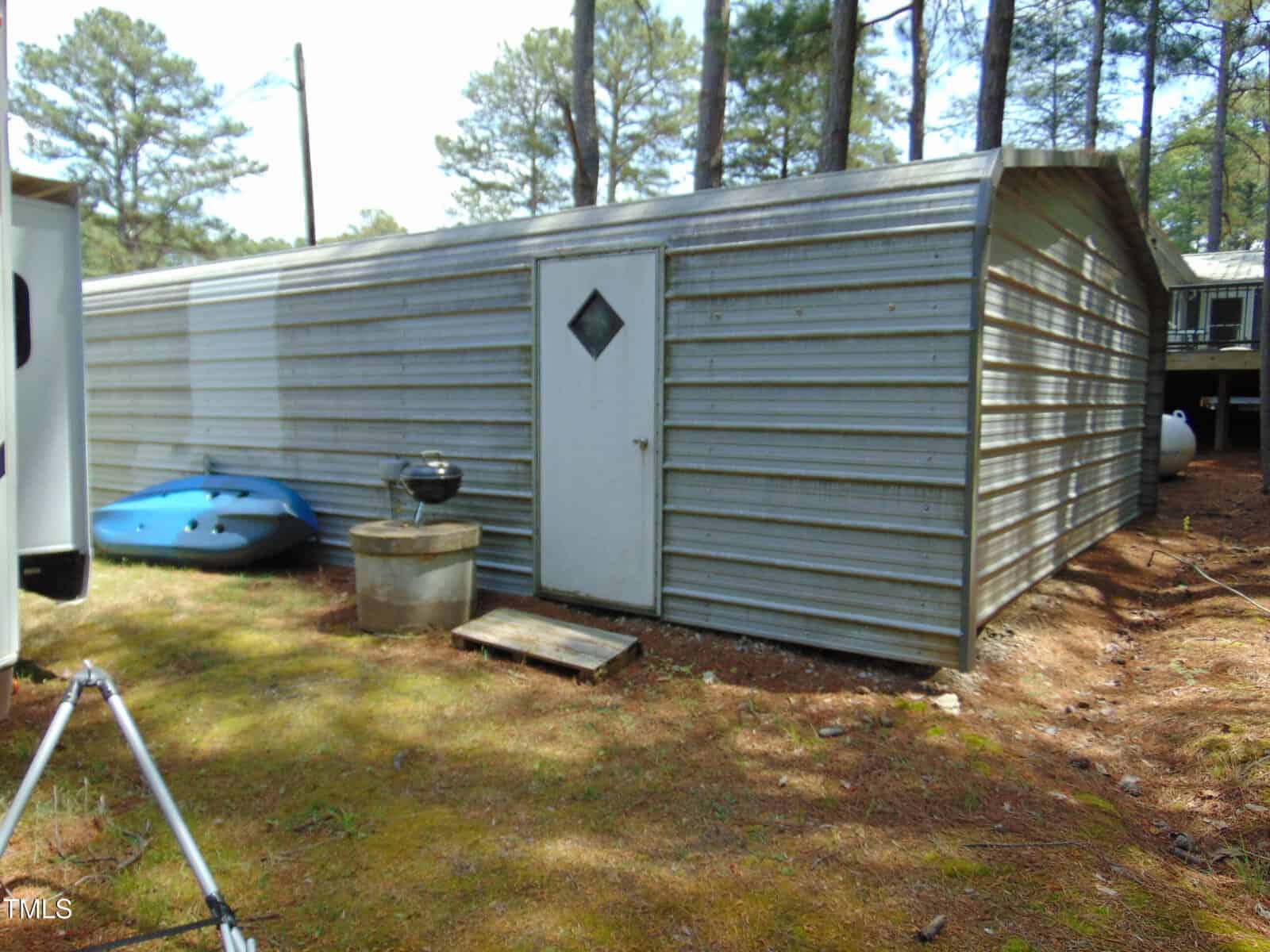 A large metal storage shed with a white door and diamond-shaped window sits on a pine-covered lot. A blue kayak, black grill, and concrete blocks are placed near the shed. Tall trees surround the area.